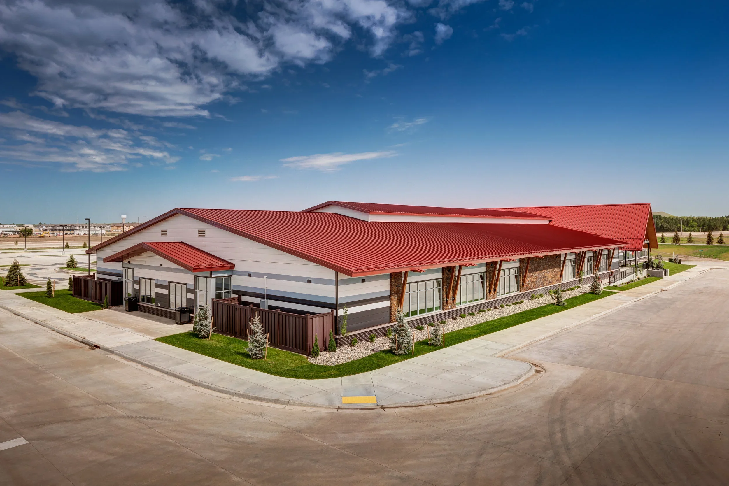 An empty commercial building with a red metal roof, white walls, large windows, and a small fenced outdoor area with trees and grass, situated in an empty parking lot under a blue sky with some clouds.
