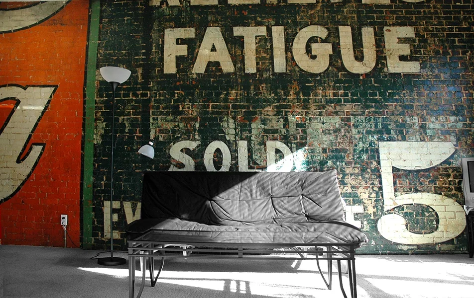 Living room with a black leather sofa, a tall floor lamp, a brick wall with large white painted letters spelling 'FATIGUE' and other words partially visible, and a TV on a stand.