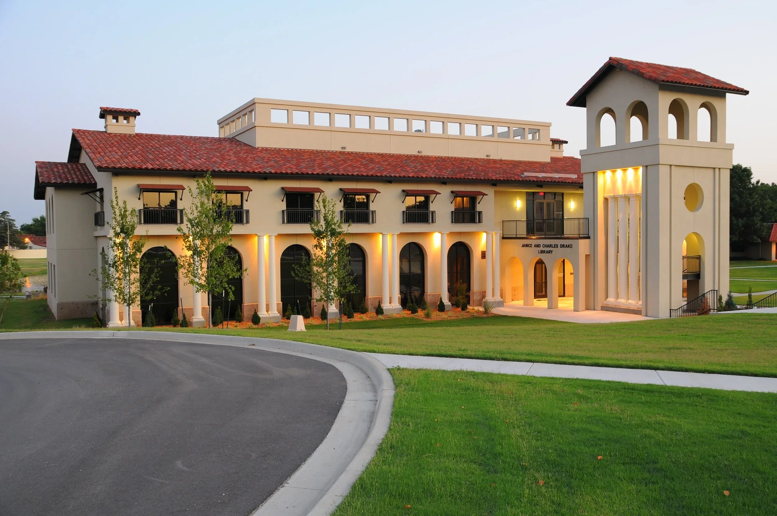 A large building with arched windows, a red roof, columns, and a tower, identified as the Janice and Charles Drake Library, illuminated at dusk with surrounding well-kept lawn and trees.