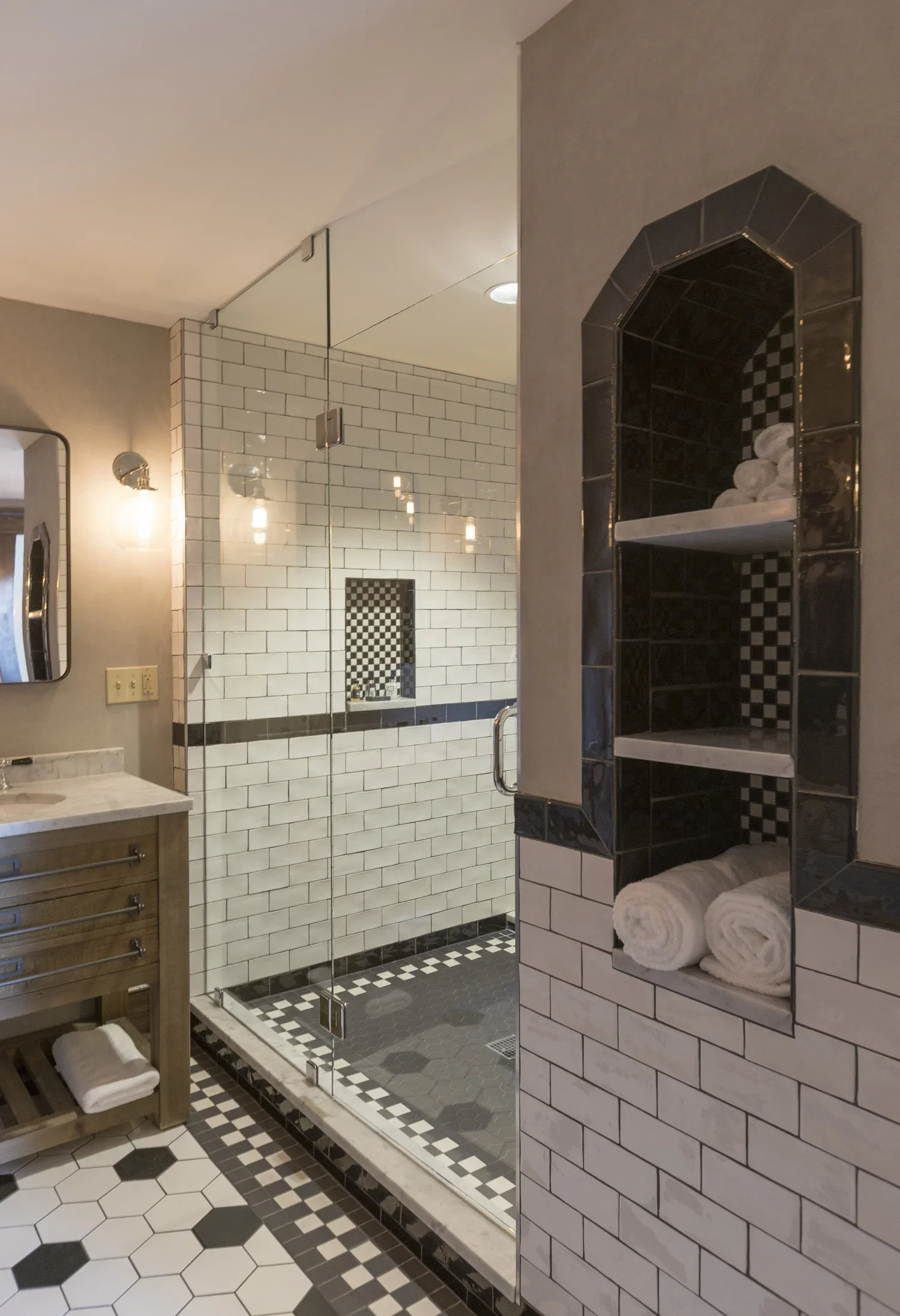 A bathroom with a glass shower enclosure, black and white tile flooring, white subway tile walls, and a small tiled nook with rolled towels and toiletries.