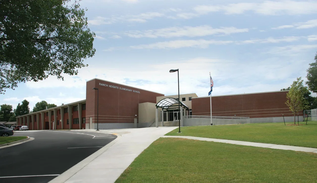 Exterior view of Ranch Heights Elementary School building with a parking lot, green lawn, and flags, under a cloudy sky.