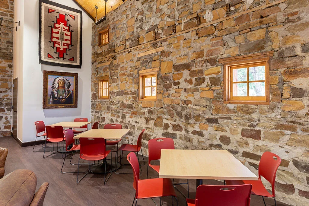 A cozy cafe interior with a rustic brick wall, small square tables with red chairs, small windows, and Native American artwork and textile wall hangings.