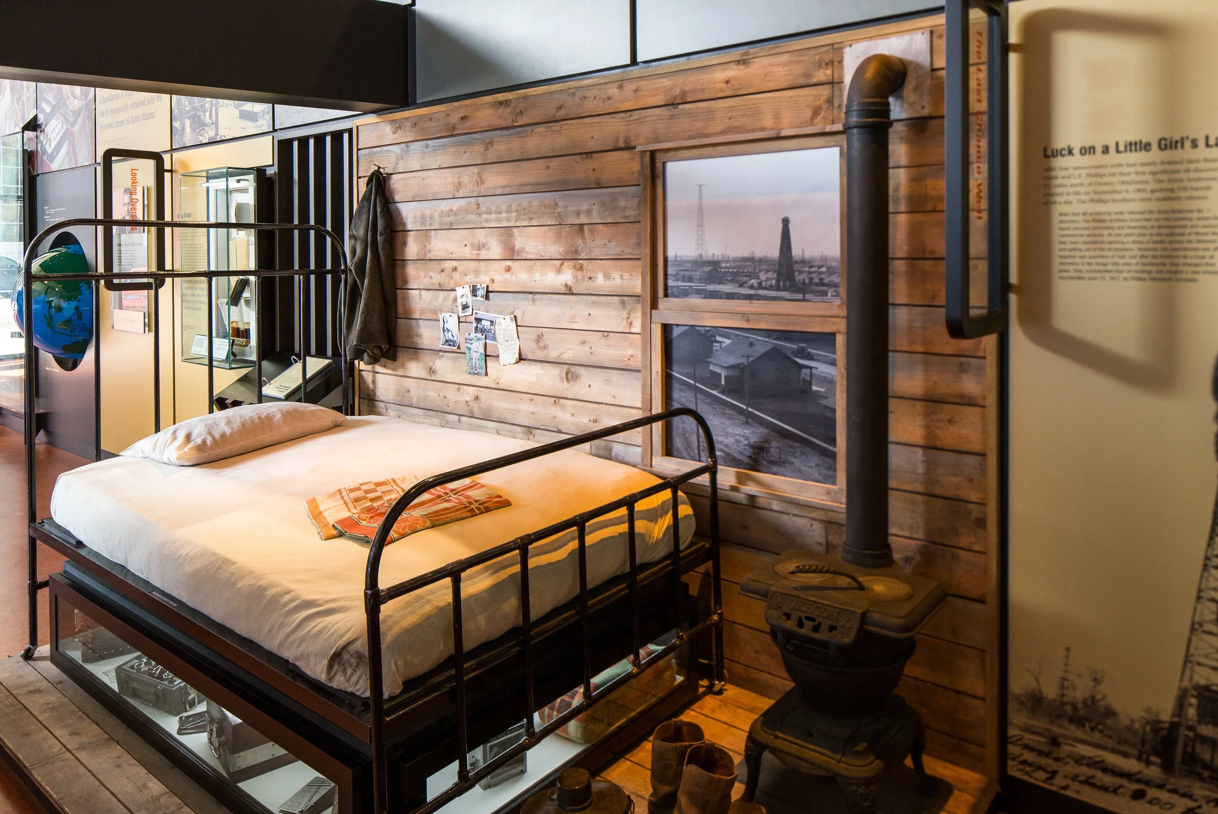 A cozy bedroom display with a black metal bed frame, white bedding, and a folded orange patterned blanket. The bed is against a wooden plank wall with a small window showing a cityscape. To the right is an old-fashioned wood stove with a tall pipe. A