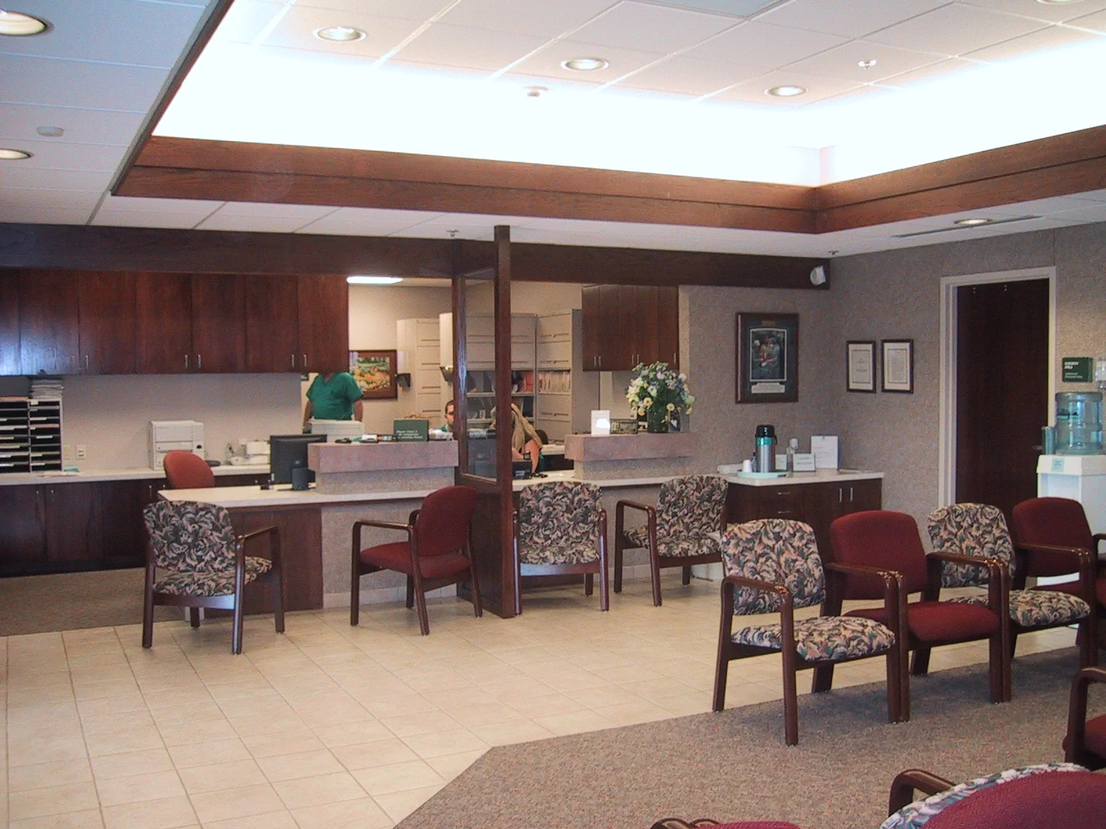 A waiting area in a medical office with rows of patterned and solid red chairs, a water cooler to the right, a reception desk with flowers, and a background area with wooden cabinets and staff, visible through an open window.