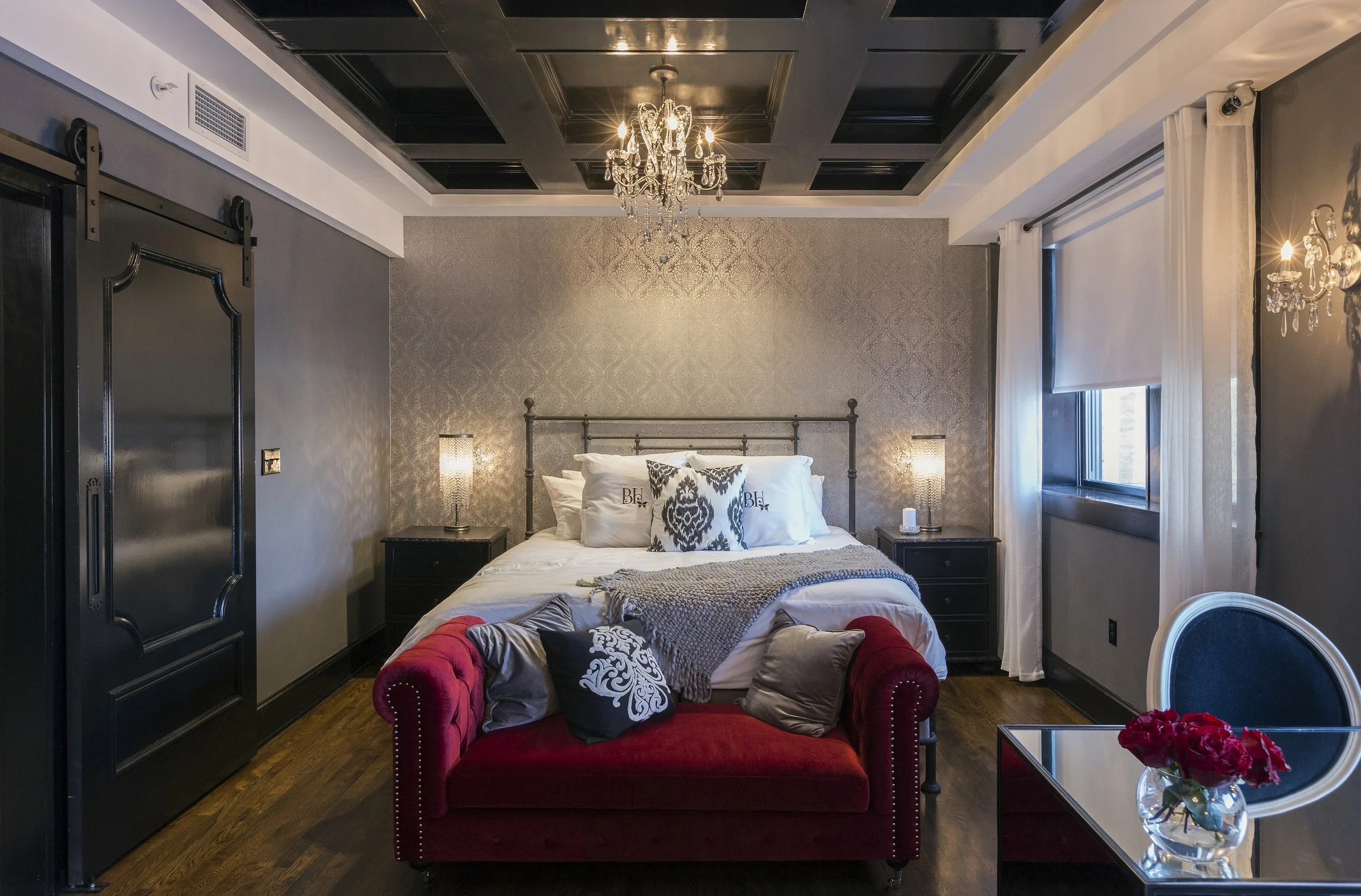A luxurious bedroom with a chandelier, a metal bed with white bedding and decorative pillows, black nightstands with lamps, a red velvet sofa at the foot of the bed with pillows, a glass-topped table with a bouquet of red flowers, a window with white