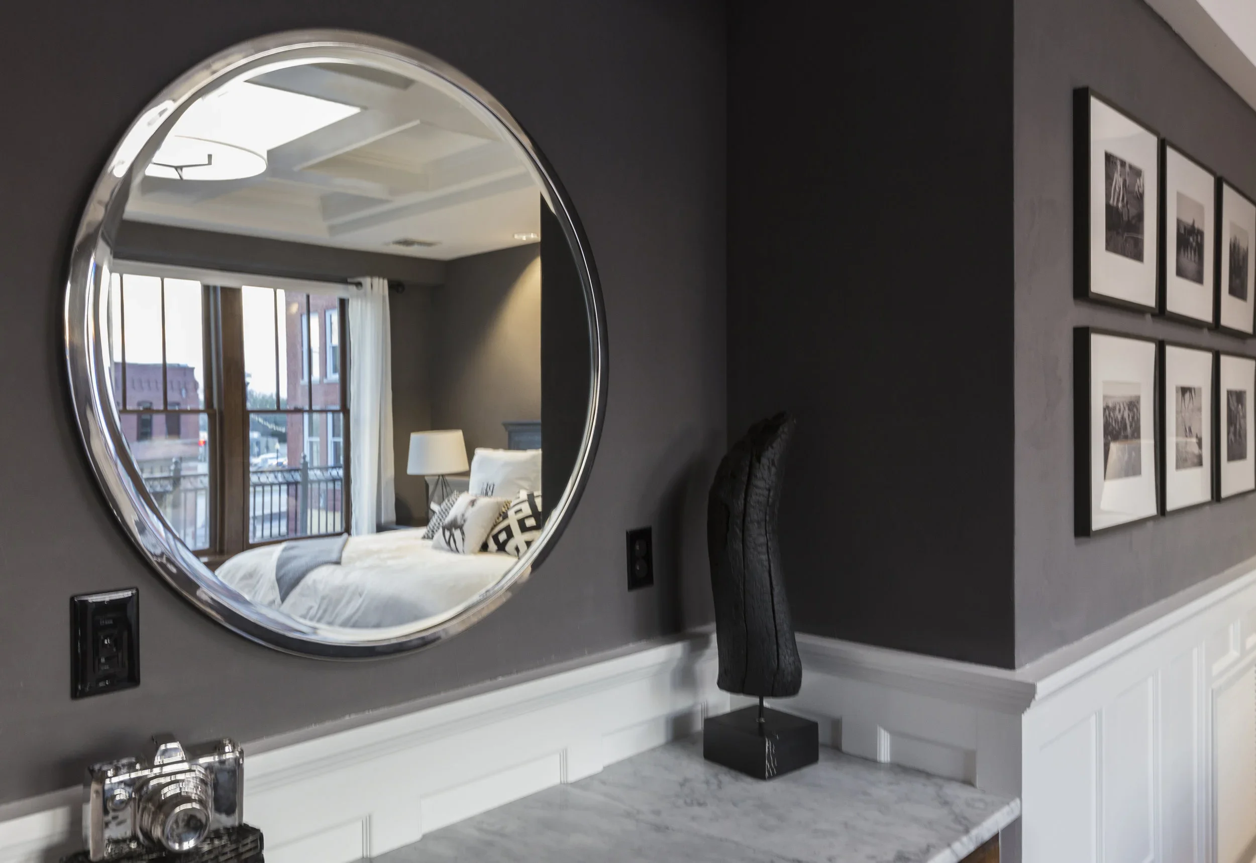 A bedroom seen through a circular mirror on a gray wall, with a bed, white curtains, framed pictures, and a sculpture on a marble surface.