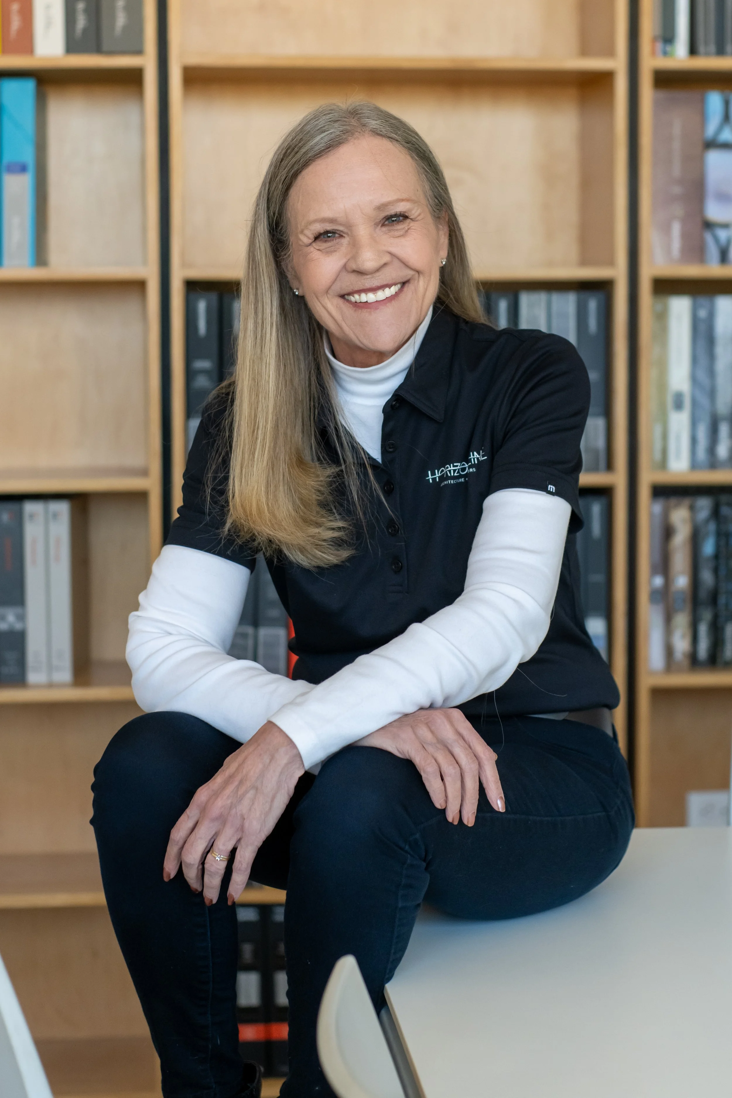 Smiling woman sitting on a table in front of wooden bookshelves filled with books and binders, wearing a black polo shirt with white long sleeves.