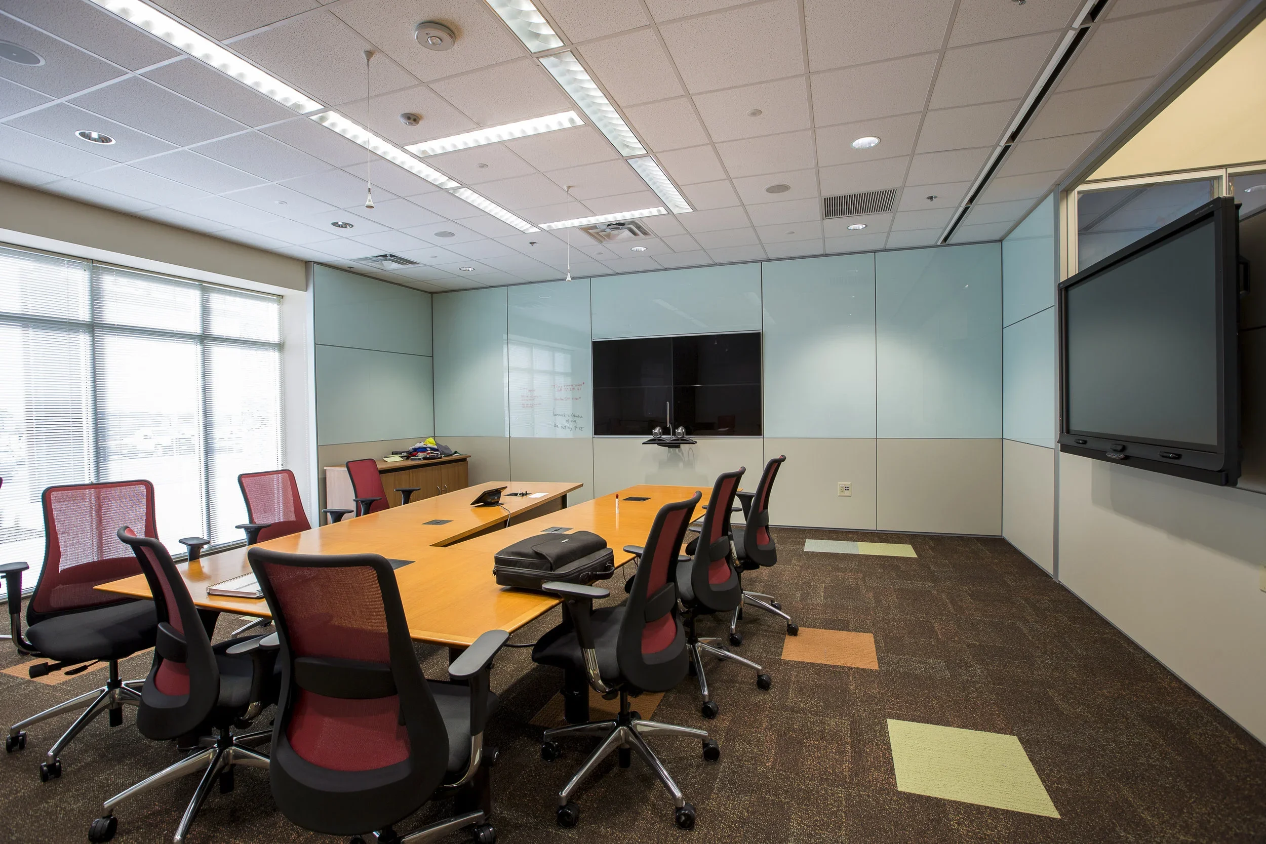 Empty conference room with a large wooden table surrounded by red and black office chairs, a wall-mounted TV screen, and a whiteboard at the front. Large windows let in natural light.