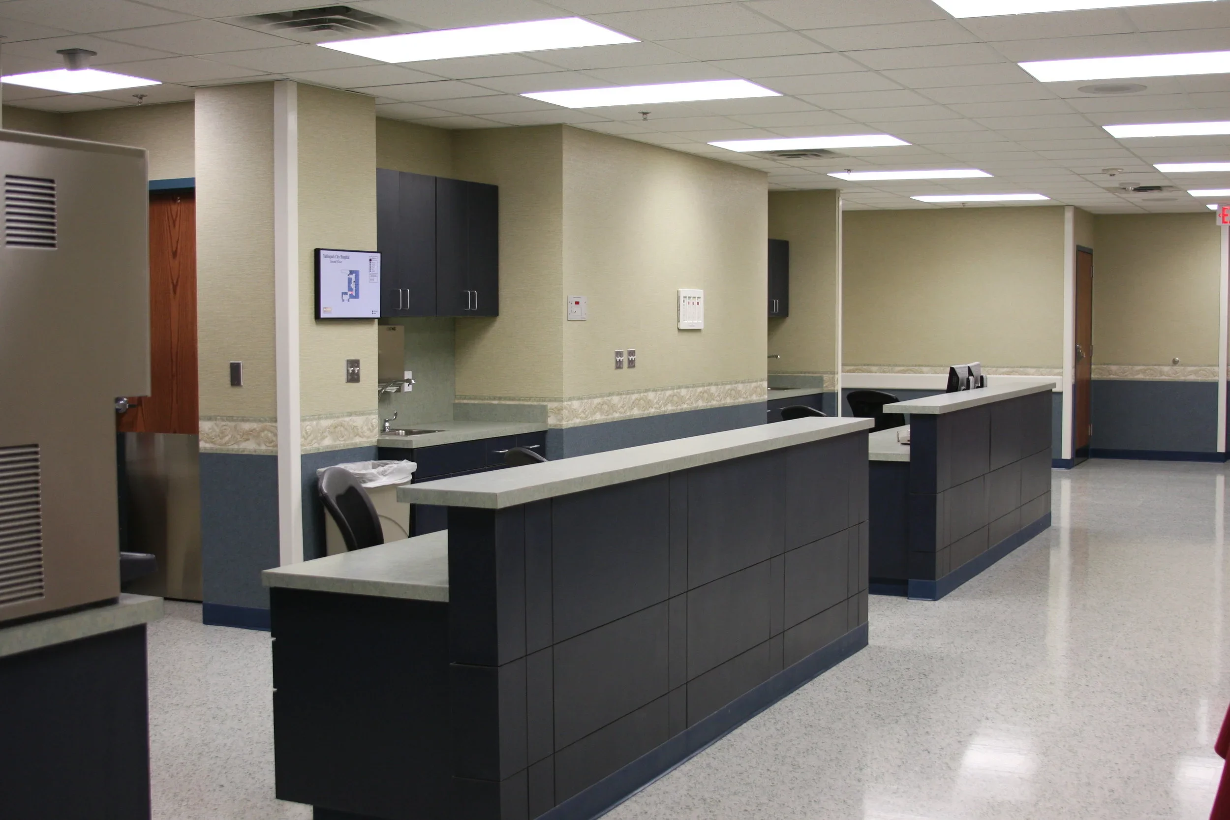Empty hospital reception area with black counters, contact chairs, and a water station, with beige and dark blue walls and ceilings with fluorescent lights.