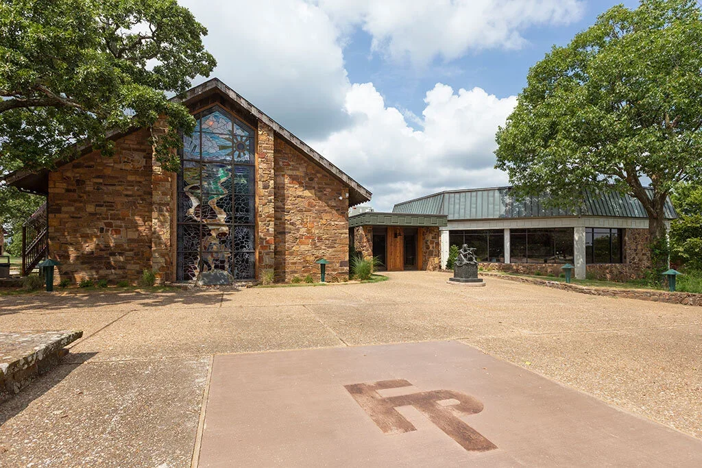 Exterior of Woolaroc Welcome Center with stone walls, a large stained glass window, and a metal roof, situated among green trees, with a concrete area in front that has a 'R' symbol etched into it.