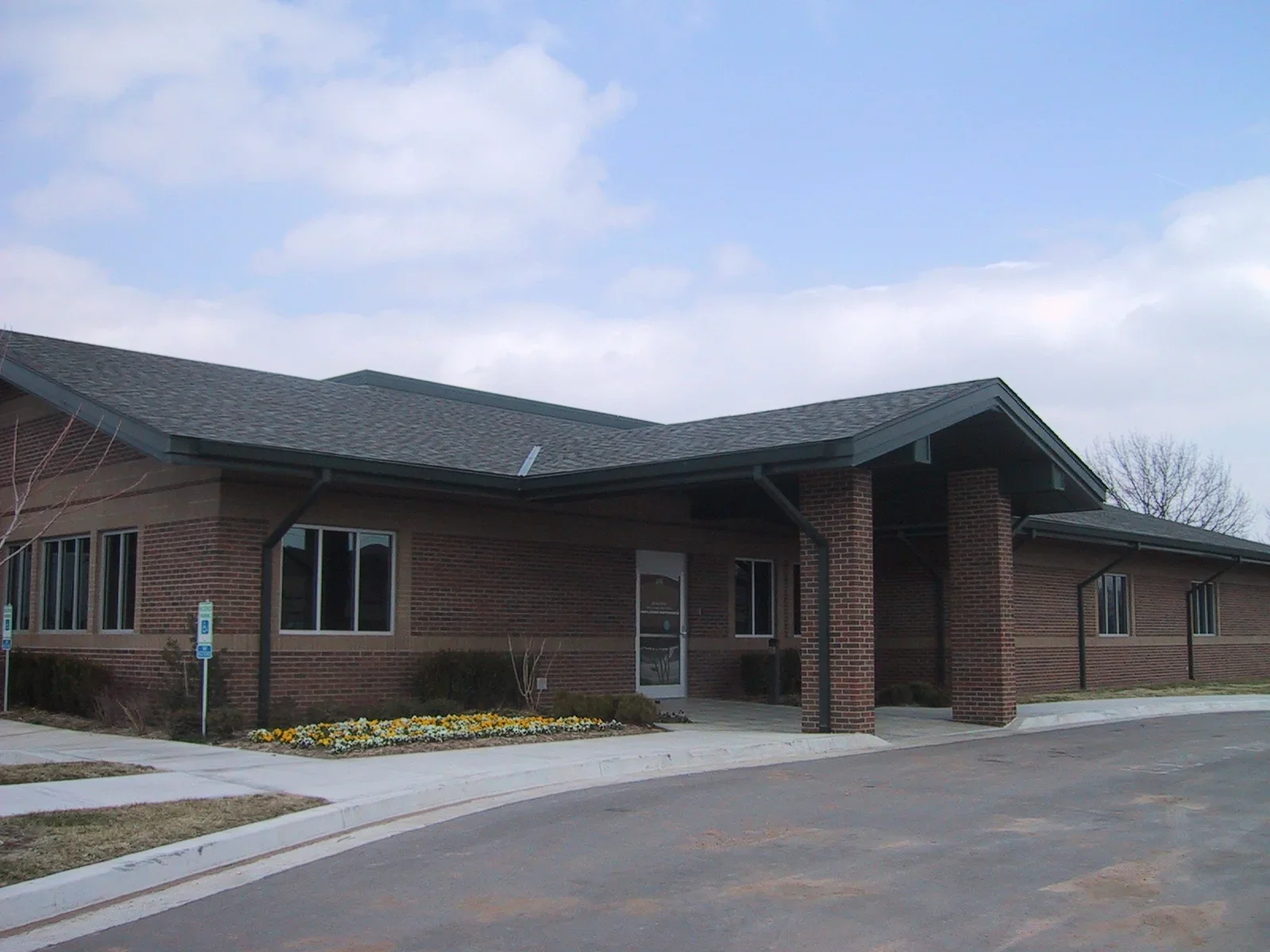 A brick building with a sloped roof, multiple windows, and a small flower bed in front, captured during daytime with a cloudy sky.