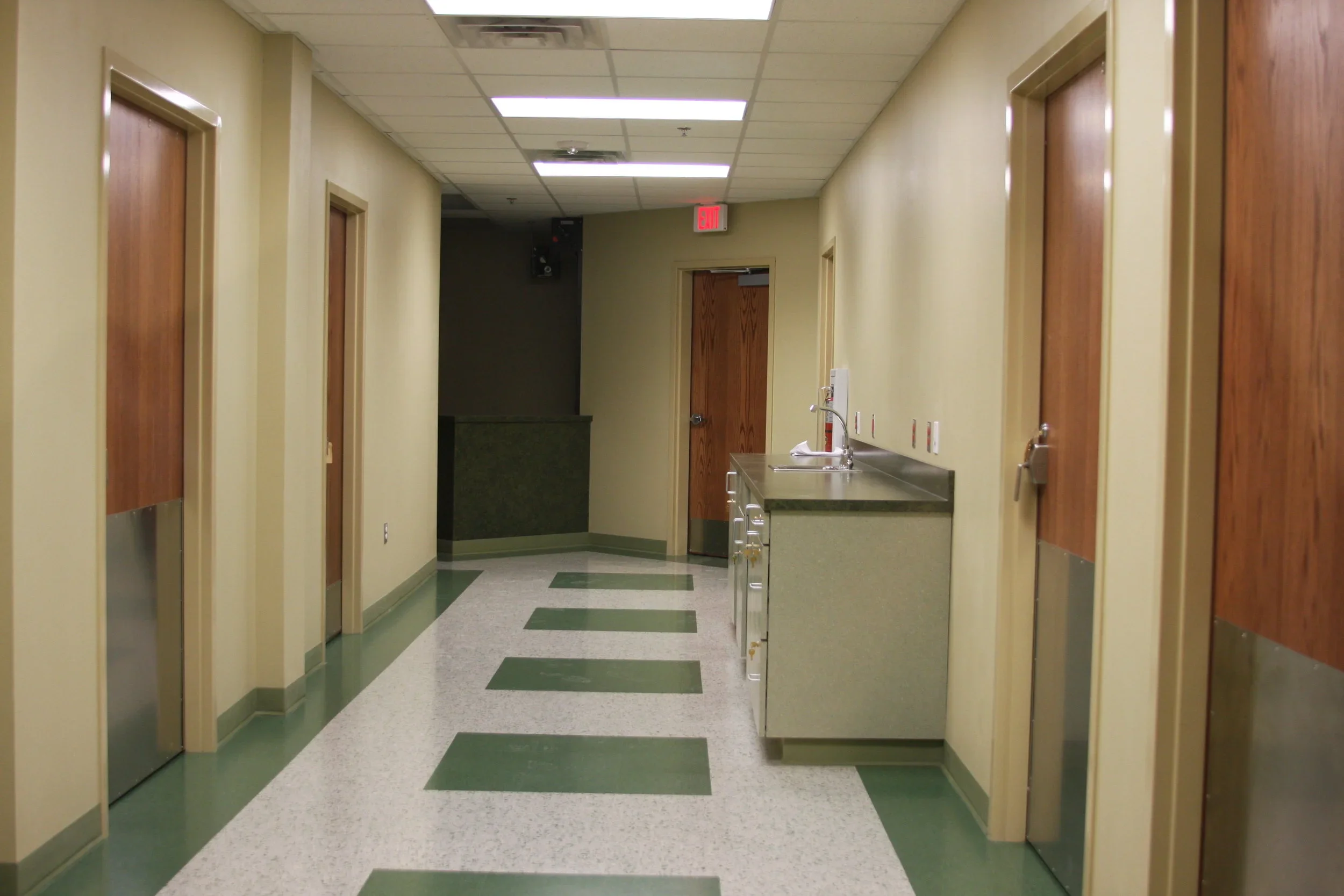 Empty hospital or clinic hallway with beige walls, green and white patterned flooring, closed wooden doors, a sink with a countertop on the right, and an exit sign above a door at the end of the hall.