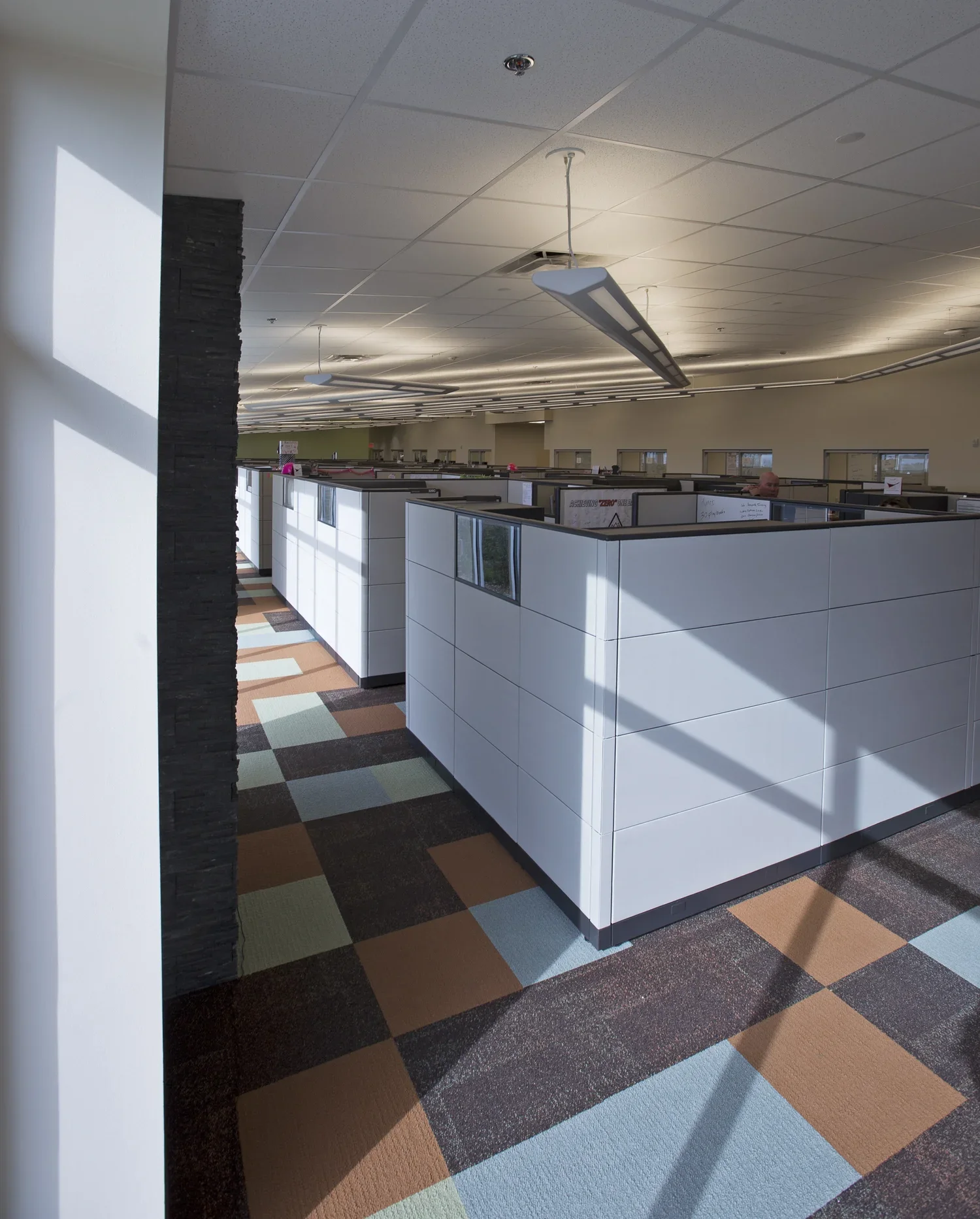 Empty office cubicles with white partitions, multicolored carpet tiles, fluorescent lighting, and ceiling vents, with sunlight casting shadows inside.