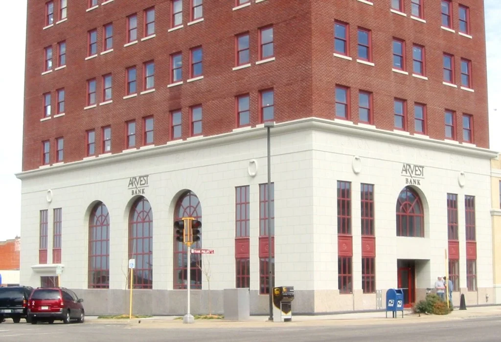 A brick and cream-colored building with arched windows houses the Arvest Bank. There are cars, street signs, and mailboxes in front of the building.