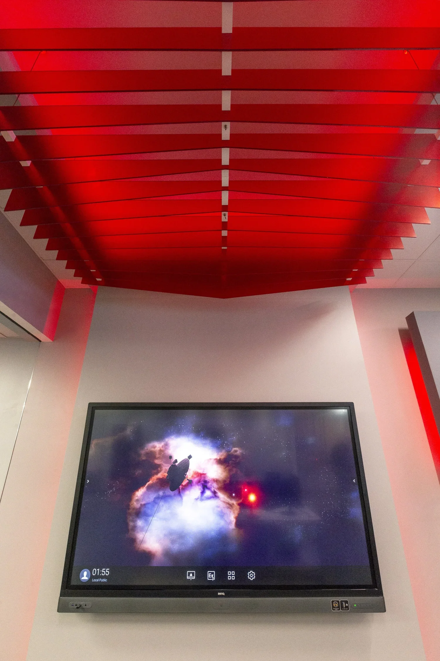 A digital display screen showing an image of a space scene with a spaceship, colorful nebulae, and cosmic clouds, mounted on a wall beneath a ceiling with red panels.