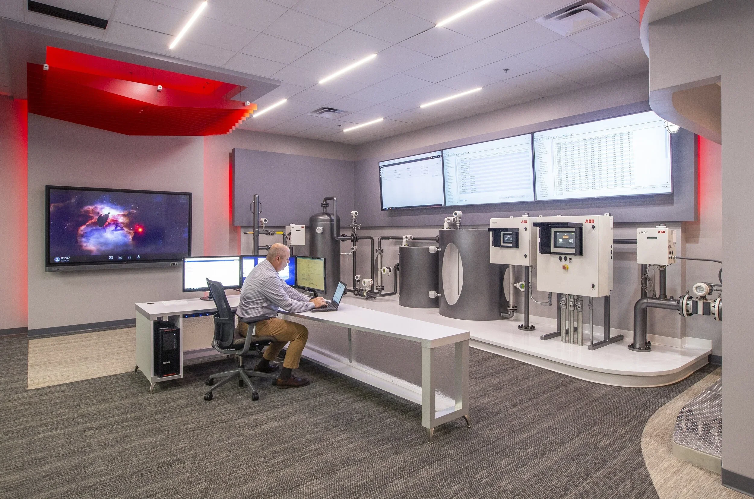 A man is sitting at a desk with multiple computer monitors, working in a high-tech control room. The room features large screens on the wall displaying data and a red-lit ceiling detail. Machinery and piping are also visible in the background.