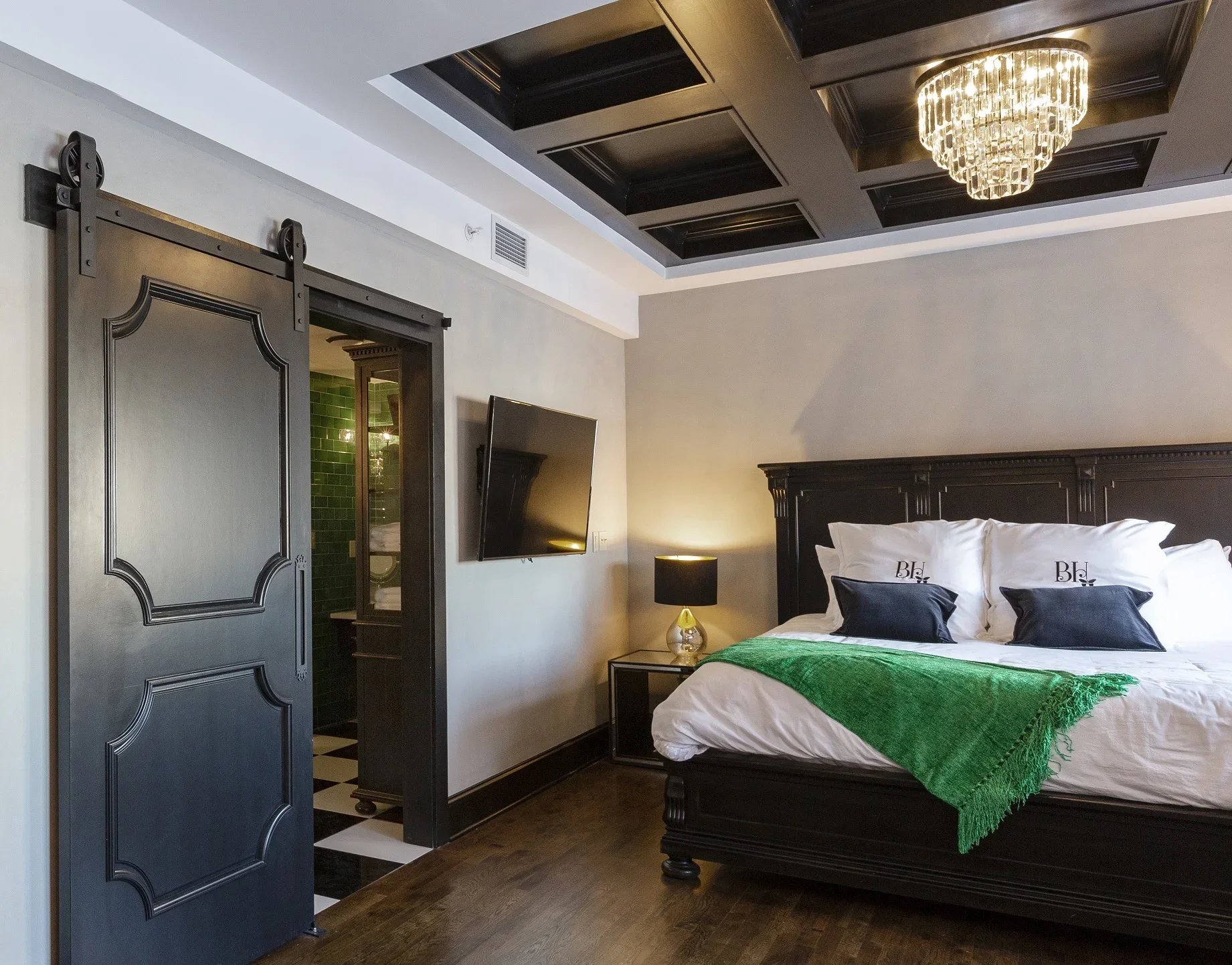 Luxury bedroom with dark wood furniture, a large bed with white linens, black pillows, green throw blanket, bedside lamp, wall-mounted TV, and a black sliding barn door leading to a bathroom with green tiles and checkered floor. Ceiling has coffered 