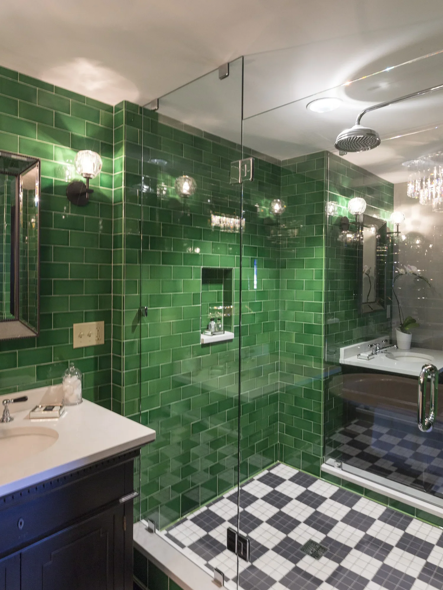 A luxurious bathroom with green tiled shower area, a glass door, and a black and white checkered floor. The bathroom features a sink with a dark cabinet, mirrors, and wall-mounted lights, along with a corner bathtub with a potted plant nearby.