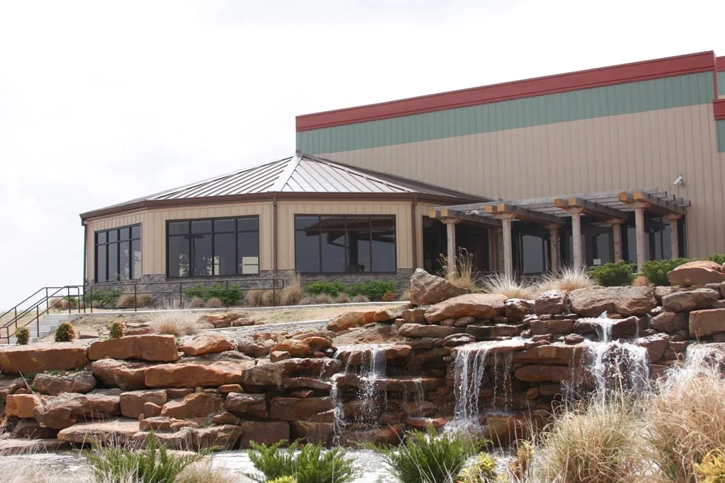Exterior view of a modern building with large windows, a sloped roof, a landscaped rocky area with water features, and a pergola, under an overcast sky.