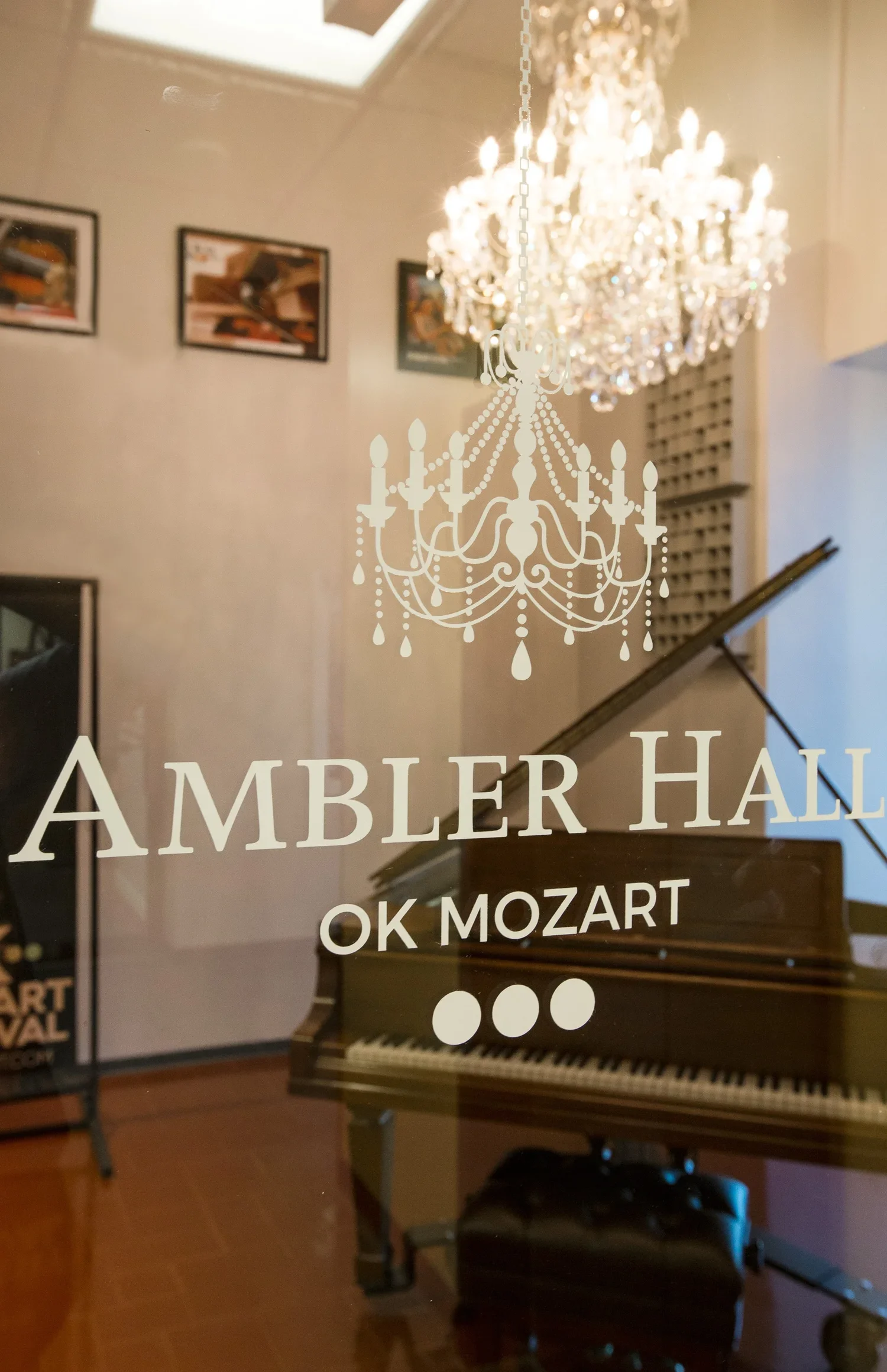 A reflection of a chandelier and a piano in a glass door with the text 'Ambler Hall OK Mozart'.