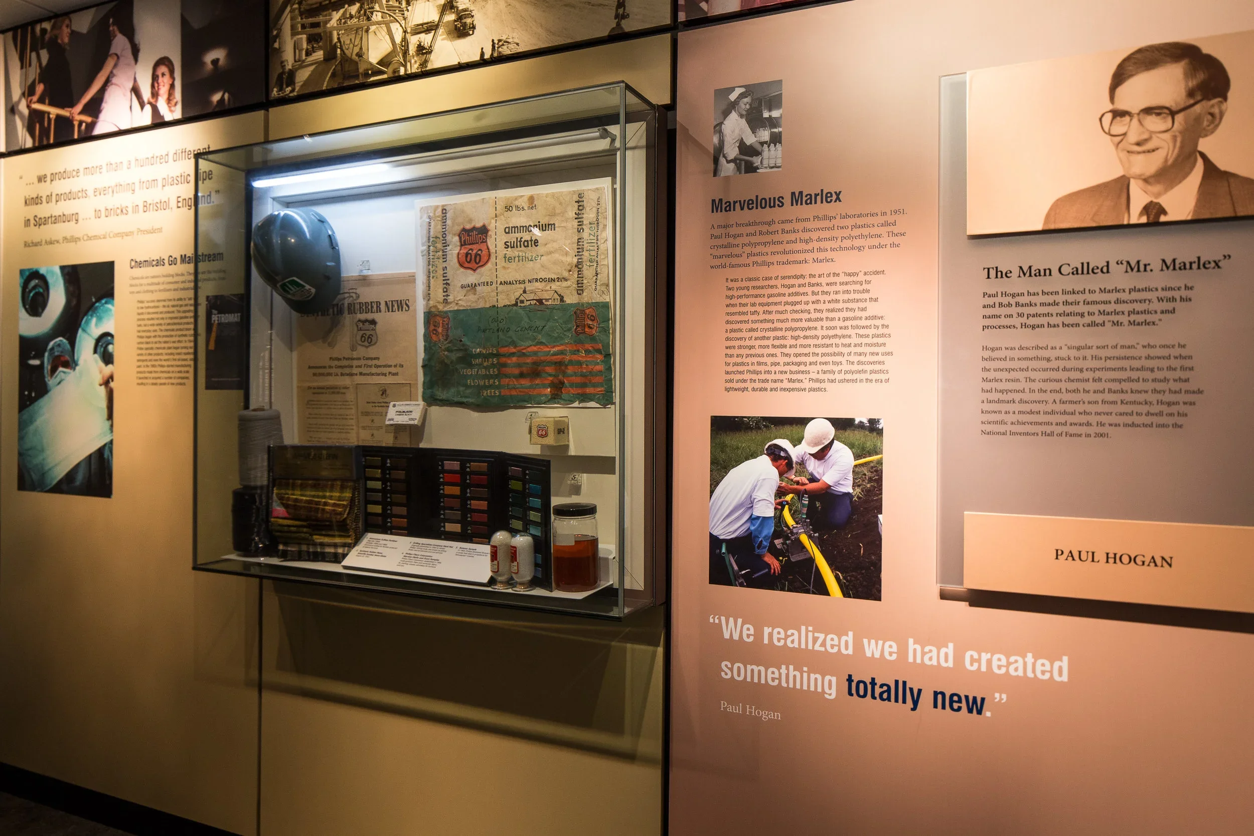Museum display with Phillips plastic manufacturing memorabilia, a helmet, color samples, and informational posters about Paul Hogan and the creation of Marlex plastic.