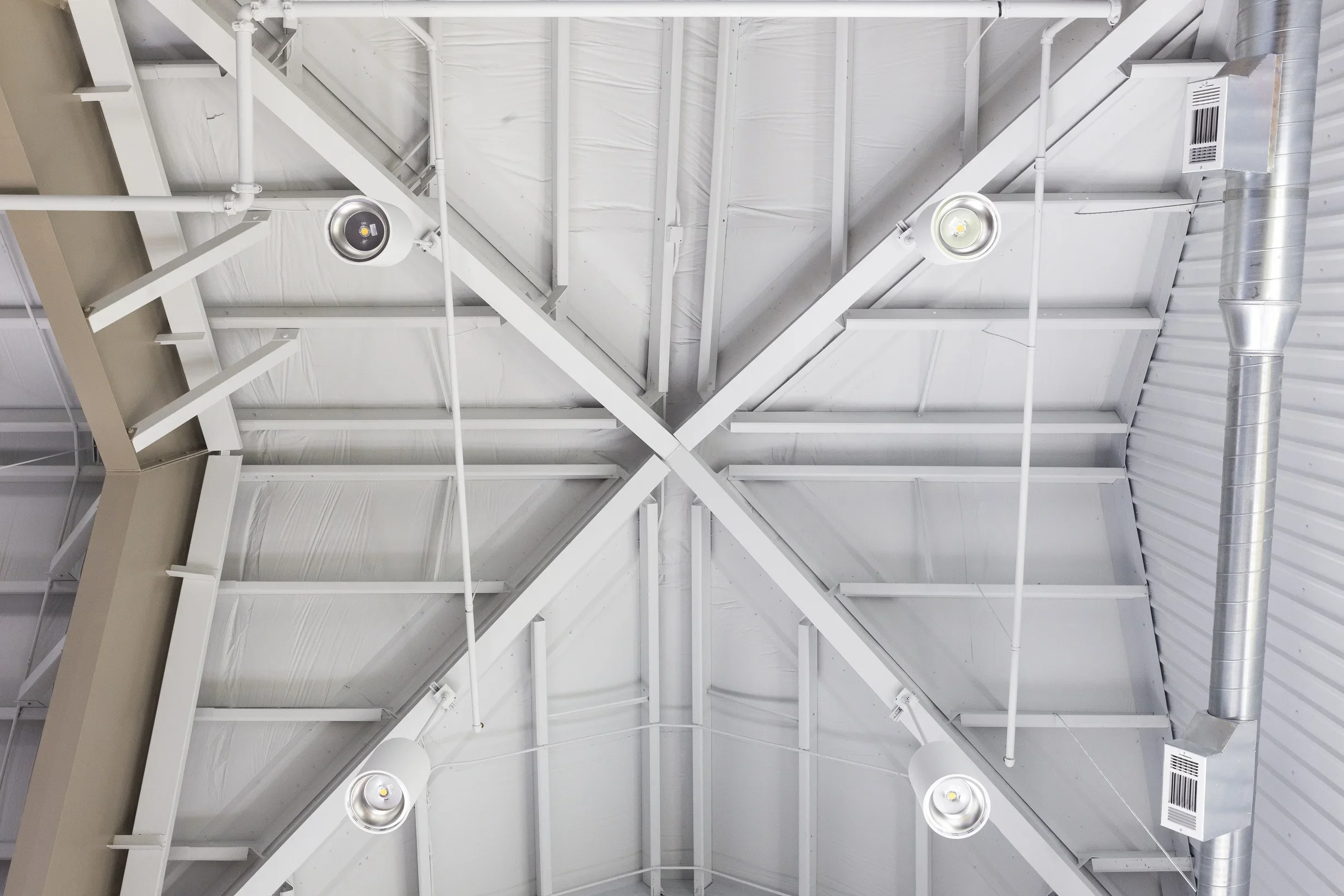 View of a white industrial ceiling with metal beams, pipes, ductwork, and four ceiling lights.