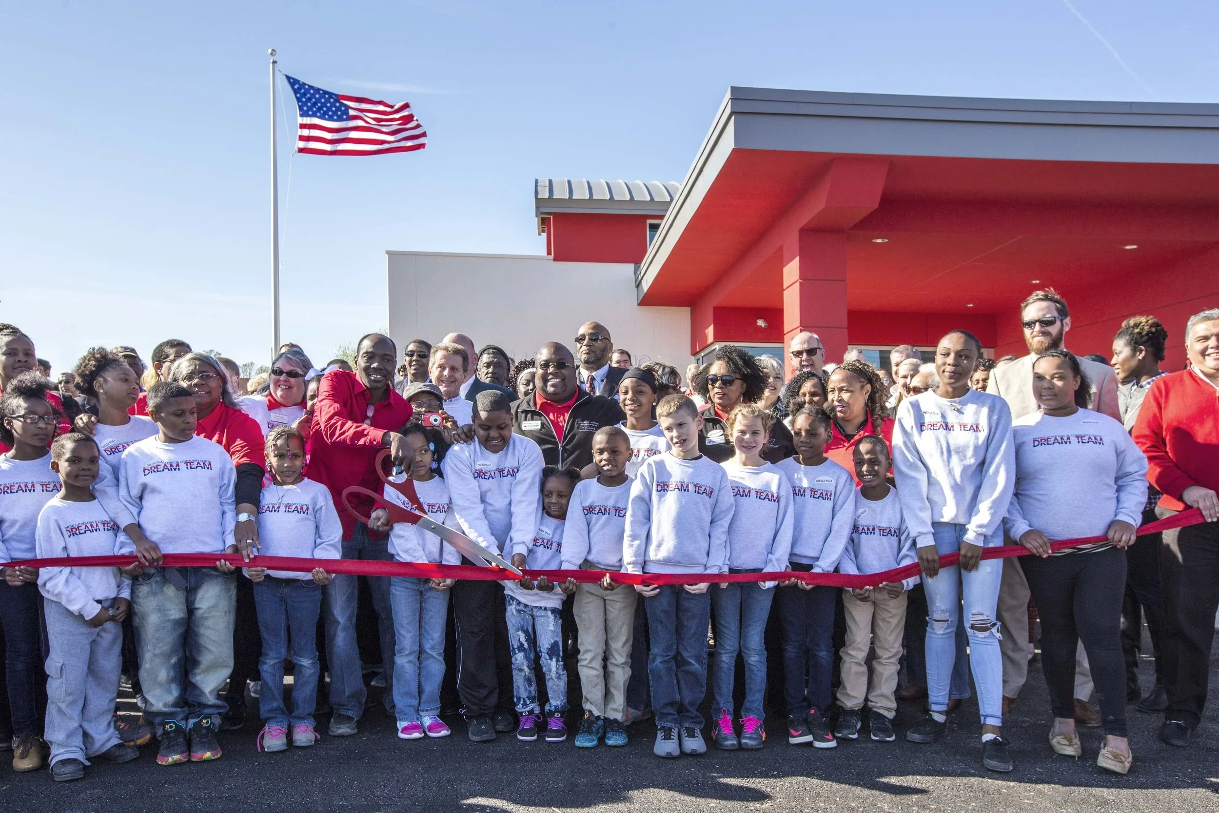 A group of children and adults participating in a ribbon-cutting ceremony outside a building with a red exterior, with an American flag flying in the background on a sunny day.