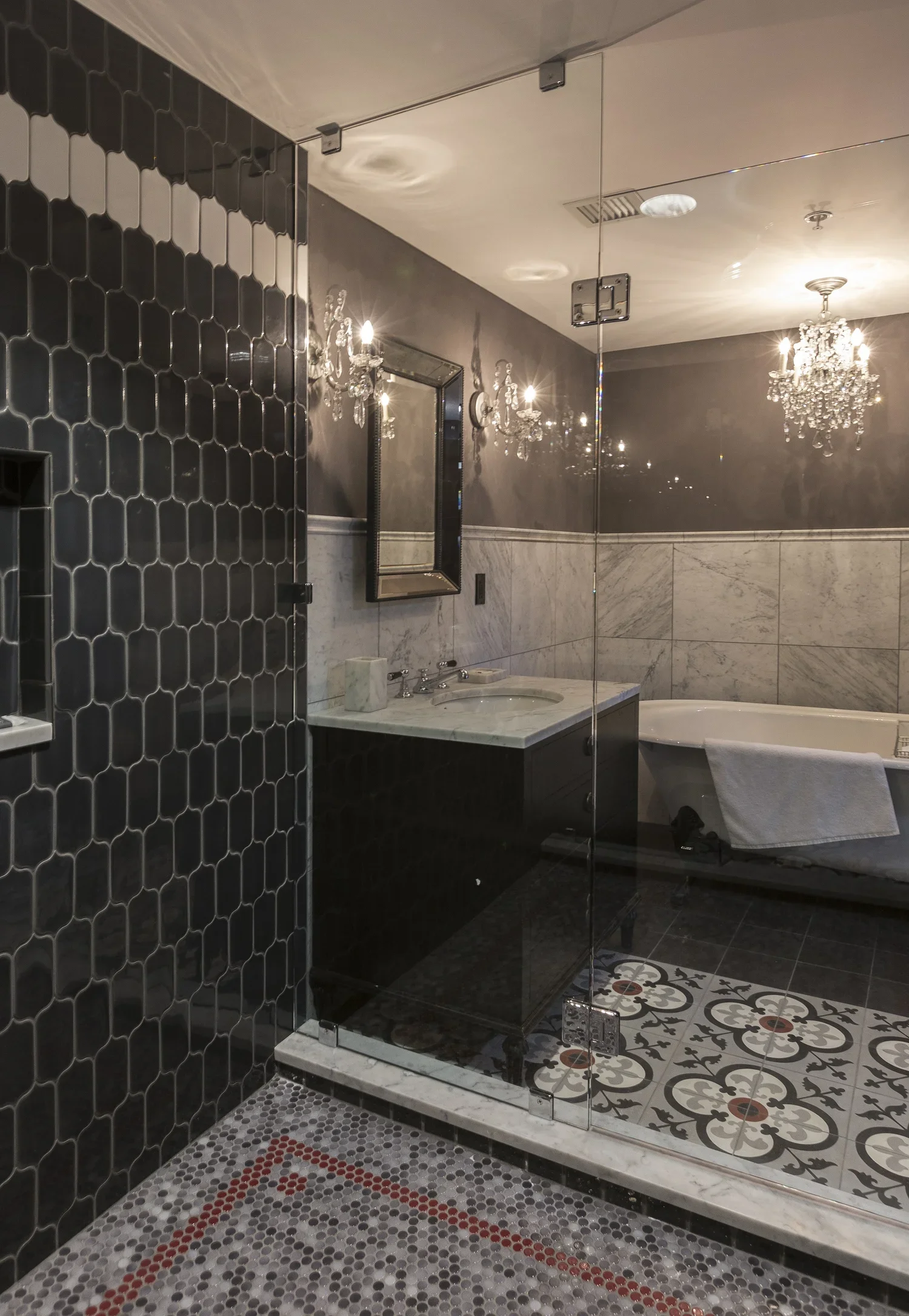 A luxurious bathroom with black and white tile floors, a glass-enclosed shower, a marble countertop with a sink, and a bathtub. The room features ornate chandeliers and wall sconces for lighting, with mirrored walls enhancing the space.