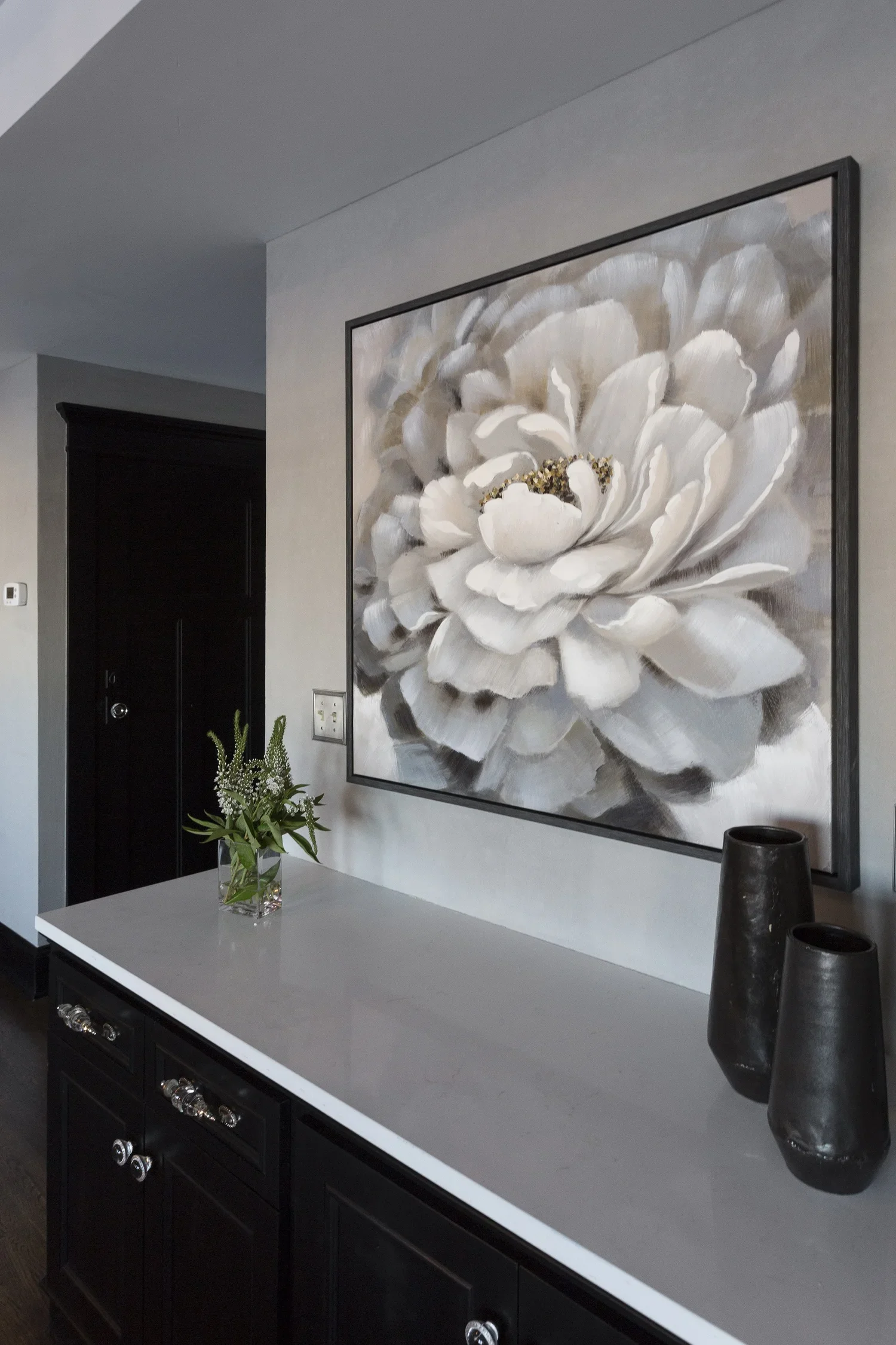Interior of a room featuring a large framed painting of a white flower, a black sideboard with silver knobs, a glass vase with green and white flowers, and three black vases on the sideboard.