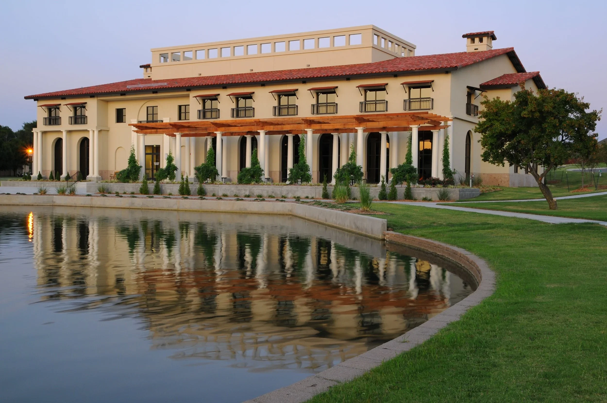 A large building with a red tile roof, white walls, and multiple balconies overlooking a pond, surrounded by a well-maintained lawn and trees.