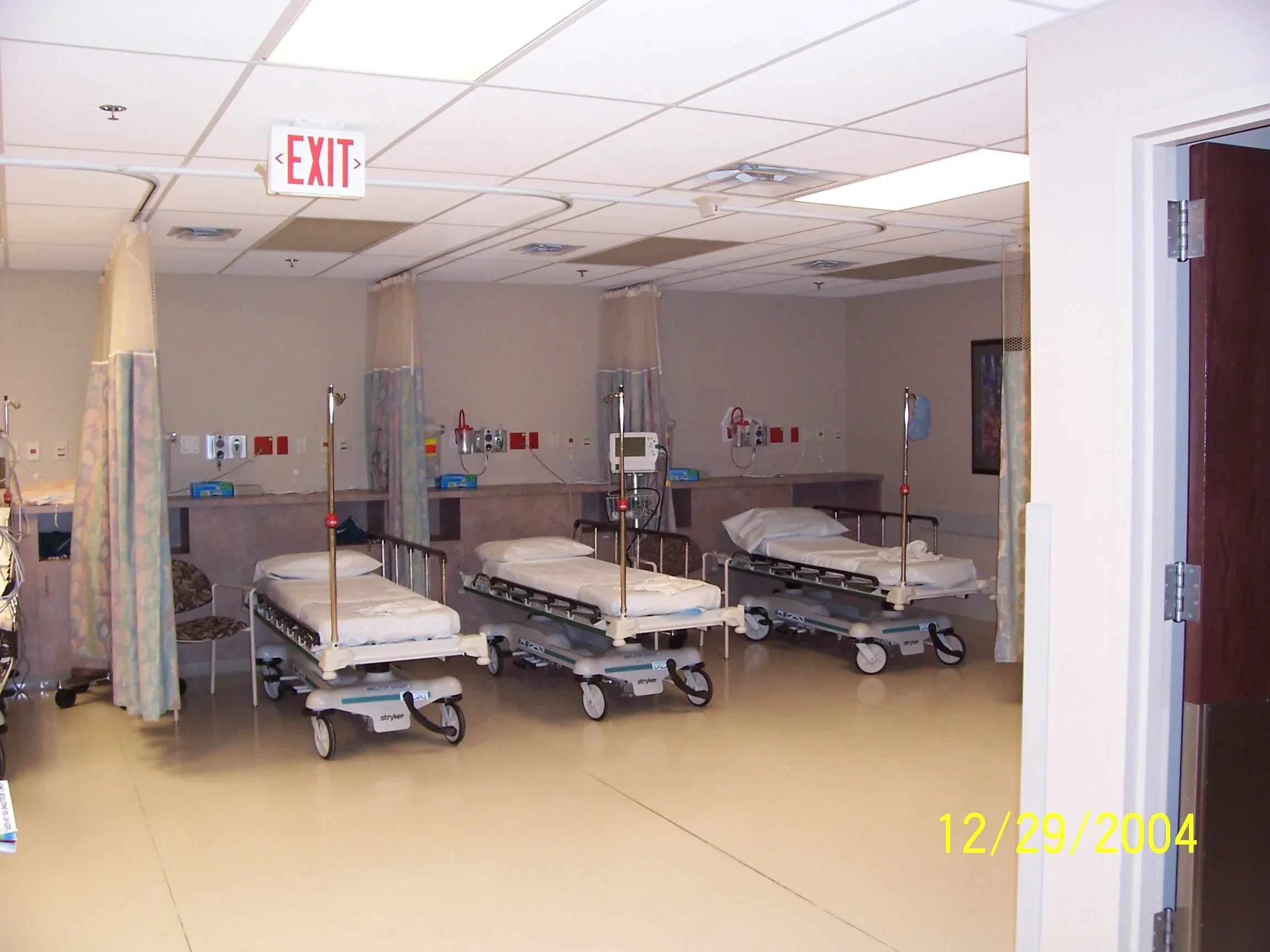 Hospital room with four empty hospital beds, each with a pillow and blanket, medical equipment on the wall, and curtains around each bed. There is an exit sign on the ceiling and the date 12/29/2004 in yellow text in the lower right corner.