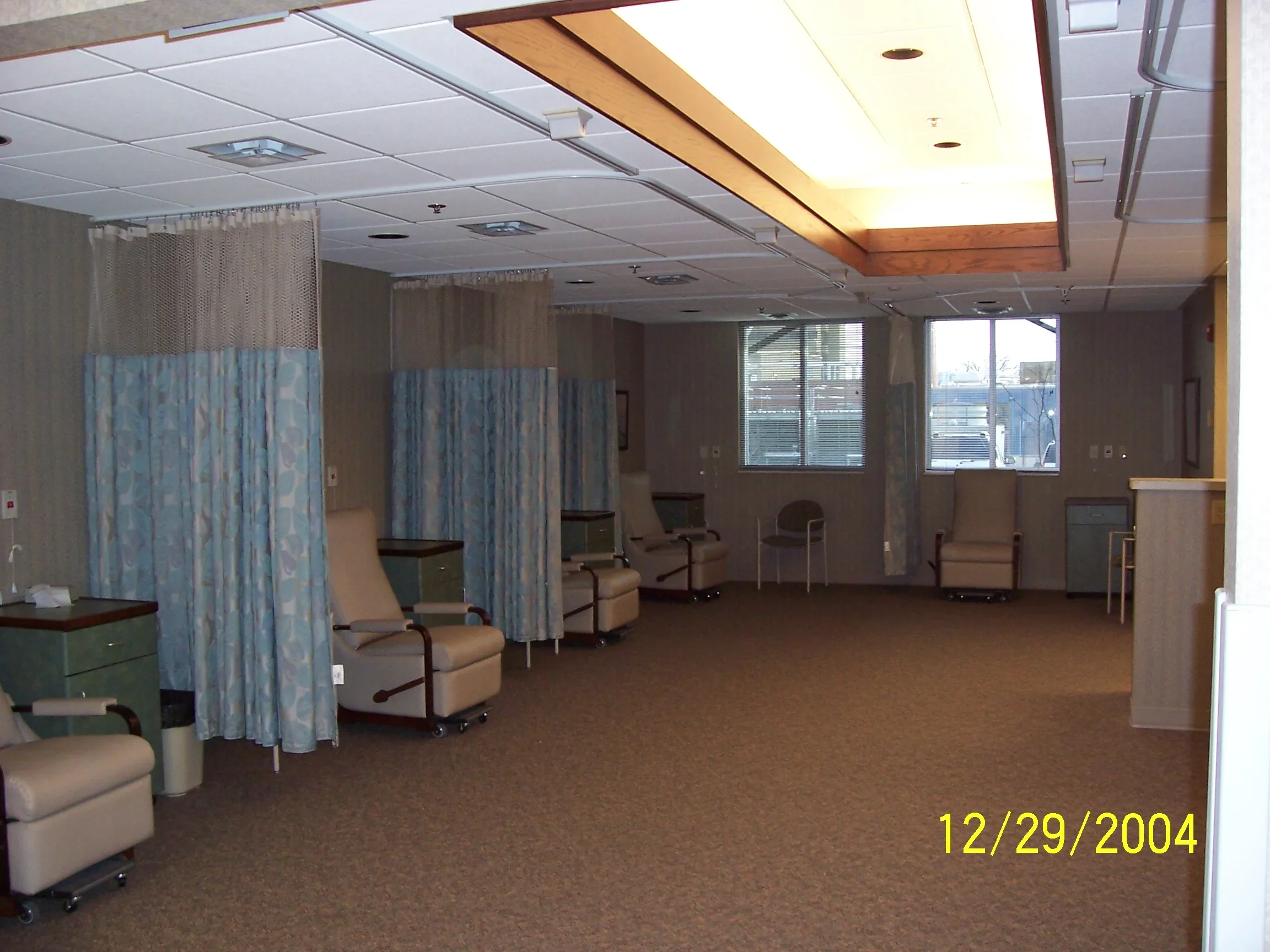 Empty hospital room with multiple patient chairs separated by curtains, and windows showing an outdoor scene.