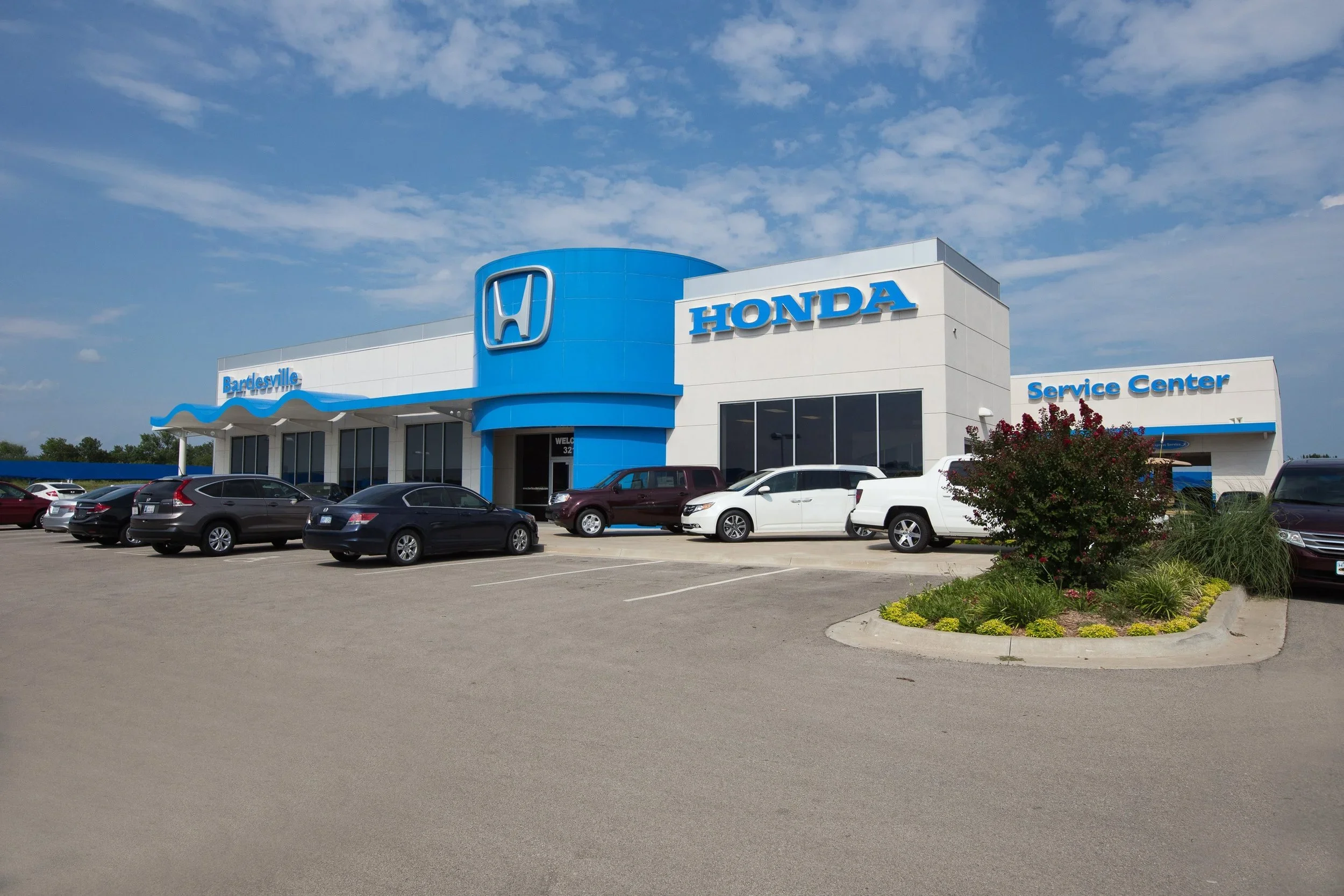 Front view of Honda dealership building with cars parked in front and a landscaped area on the right, under a partly cloudy sky.