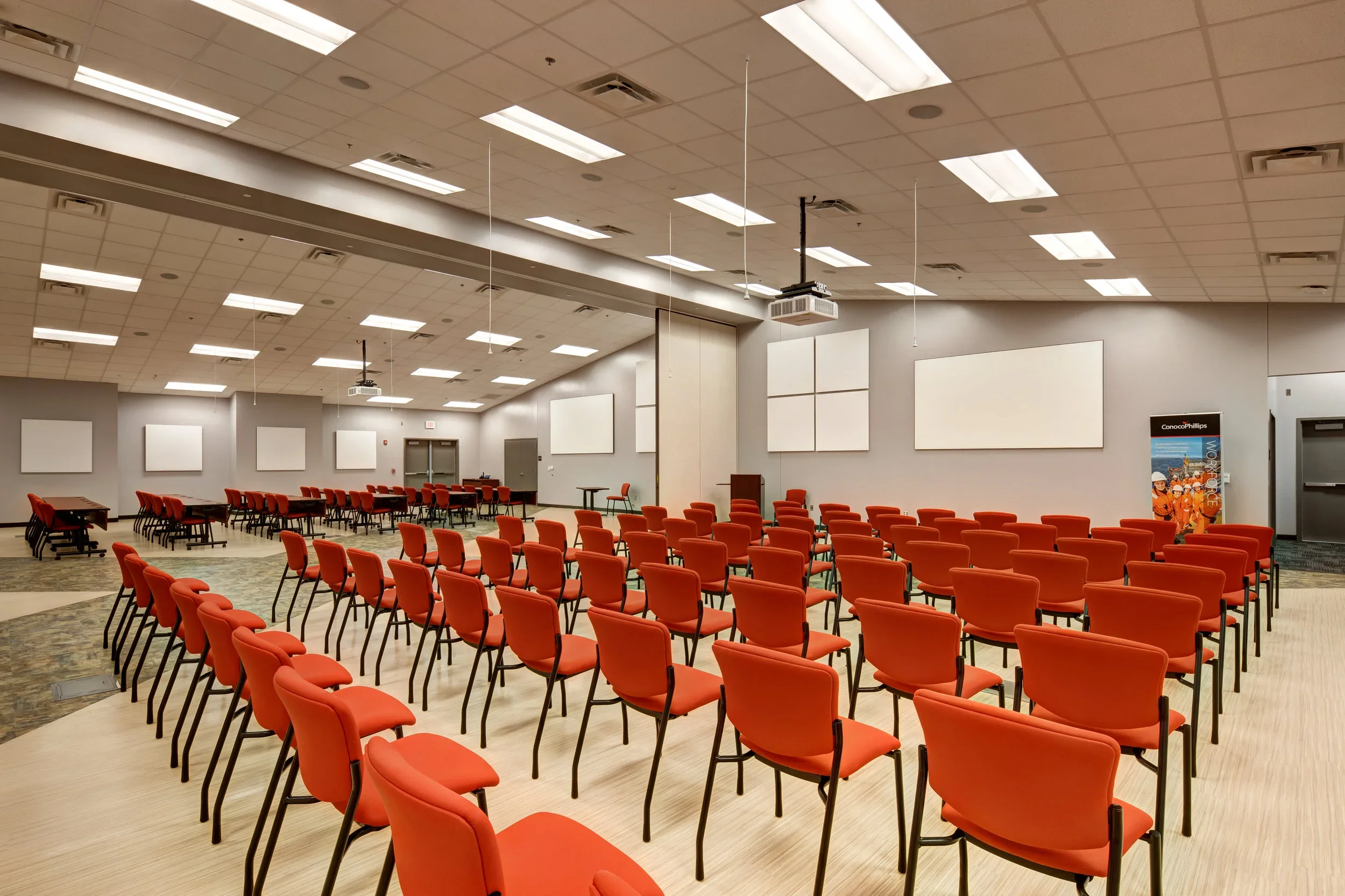 Empty conference room with rows of red chairs facing a stage with a podium and large white projection screens on the gray walls.