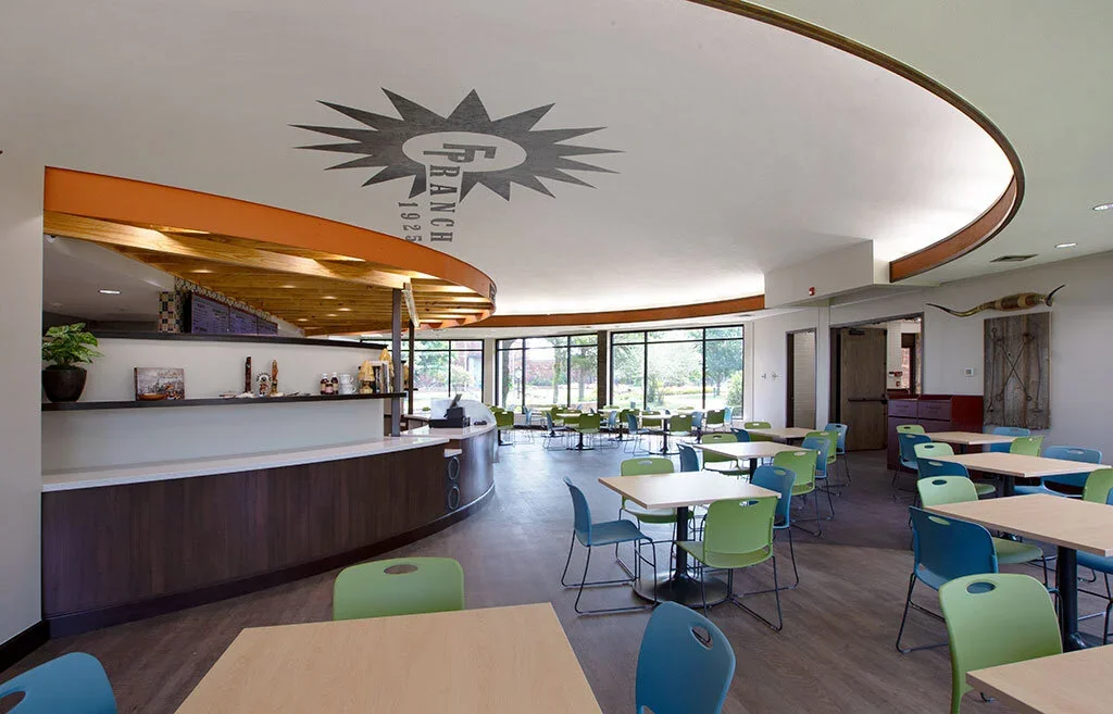 Modern restaurant or cafe with light-colored tables, blue and green chairs, large windows, and a curved ceiling with a logo. The space is bright with natural light and has a mix of seating arrangements.