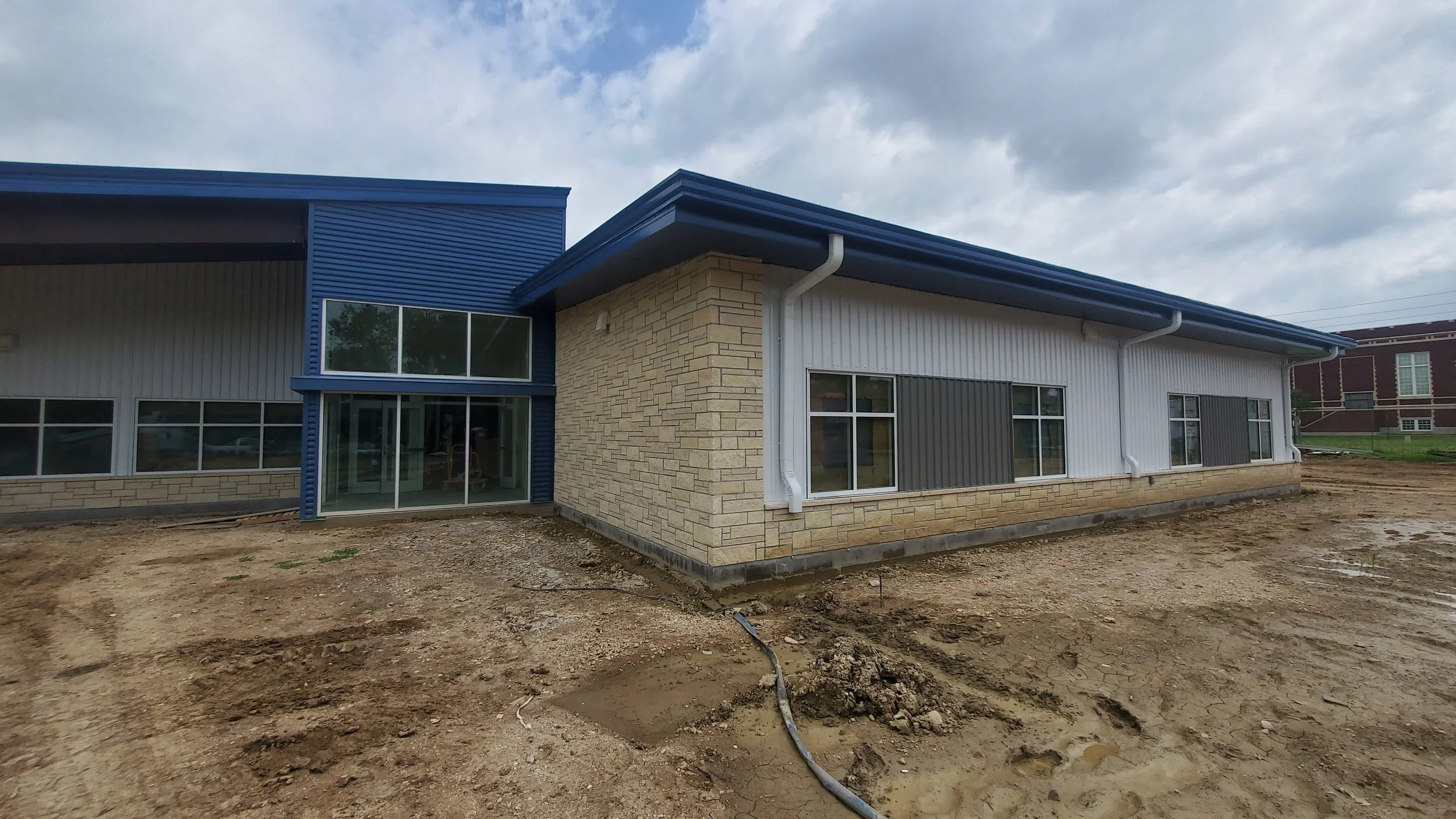 Under construction building with beige brick and blue siding exterior, large windows, and exposed ground in front.