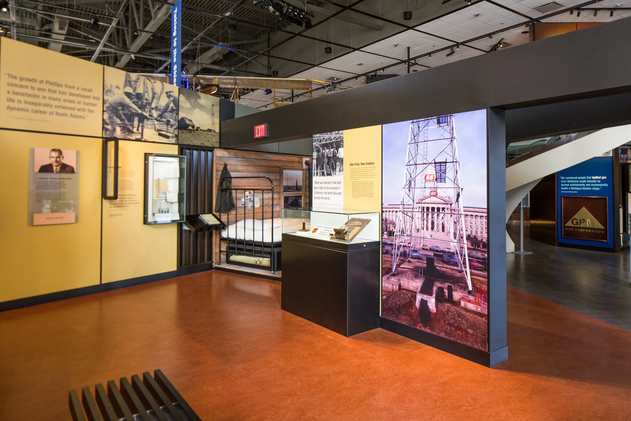 An indoor museum exhibit featuring historical displays, a bed, blue informational panels, and a large photograph of a tower with signs, with additional exhibits visible in the background.