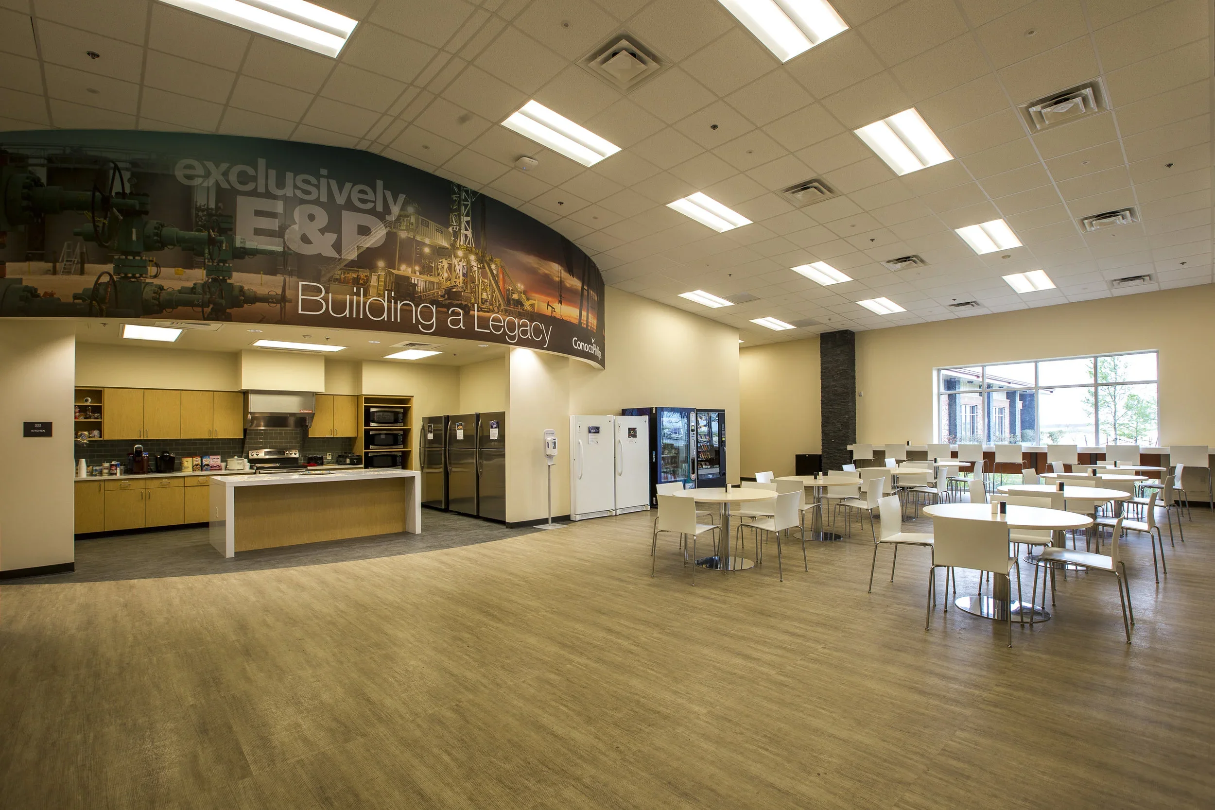 A spacious break room in an office building with a kitchenette, vending machines, and round tables with chairs for employees to sit and eat. Large windows provide natural light.