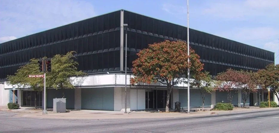 A modern, black building with large windows, trees with fall foliage, and a street corner with traffic signals.
