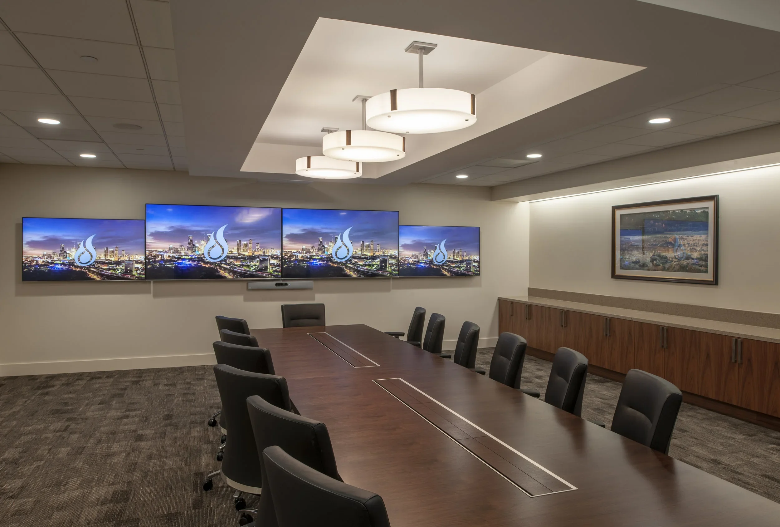 A modern conference room with a large wooden table surrounded by black chairs, three large screens displaying a city skyline at sunset with a logo, a framed landscape painting, and ceiling lights.