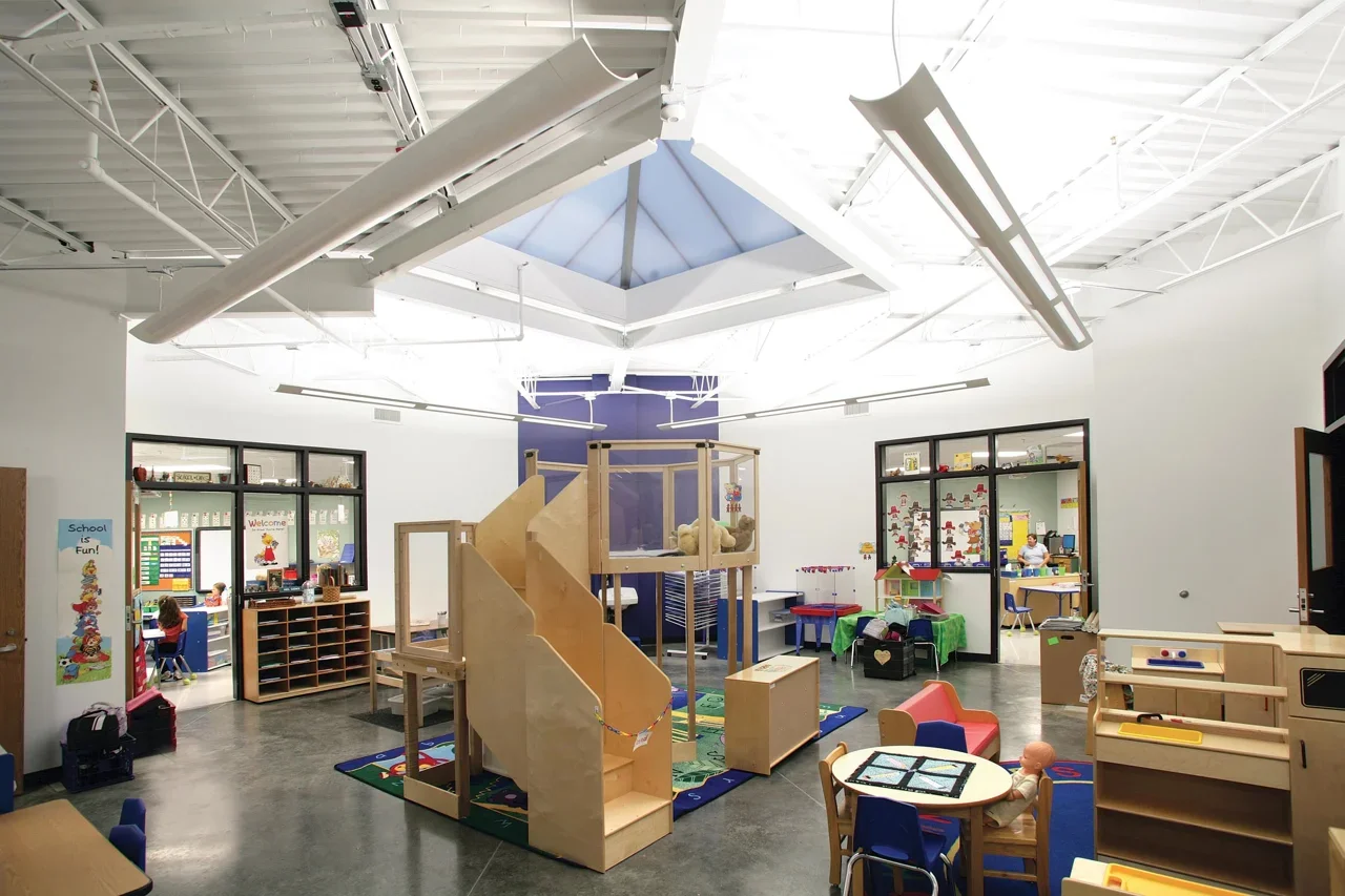 A spacious indoor children's playroom with wooden play structures, including a slide, surrounded by colorful small chairs and tables, and a play area rug. The room has ceiling lights, large windows, and doorways leading to classrooms or activity room