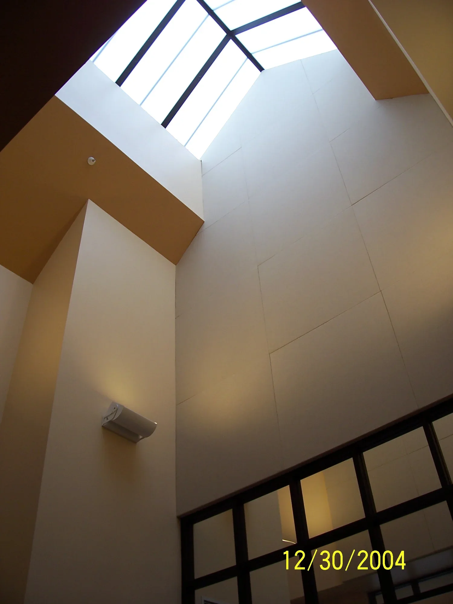 Interior shot of a modern building's atrium with high ceilings, large skylight, beige and white walls, and a section of black-framed glass windows at the bottom. 