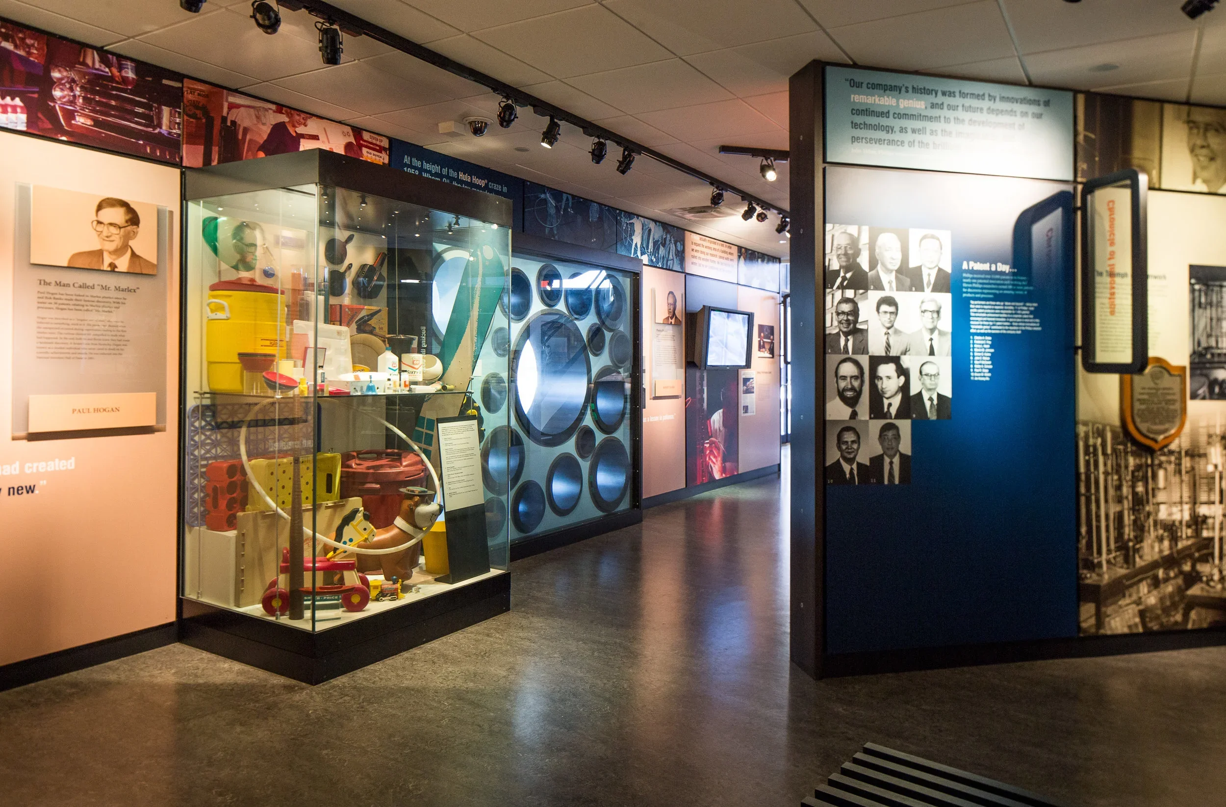 A museum exhibit with glass display case showcasing vintage items, informational panels, and photographs of people, with dim lighting and a polished floor.