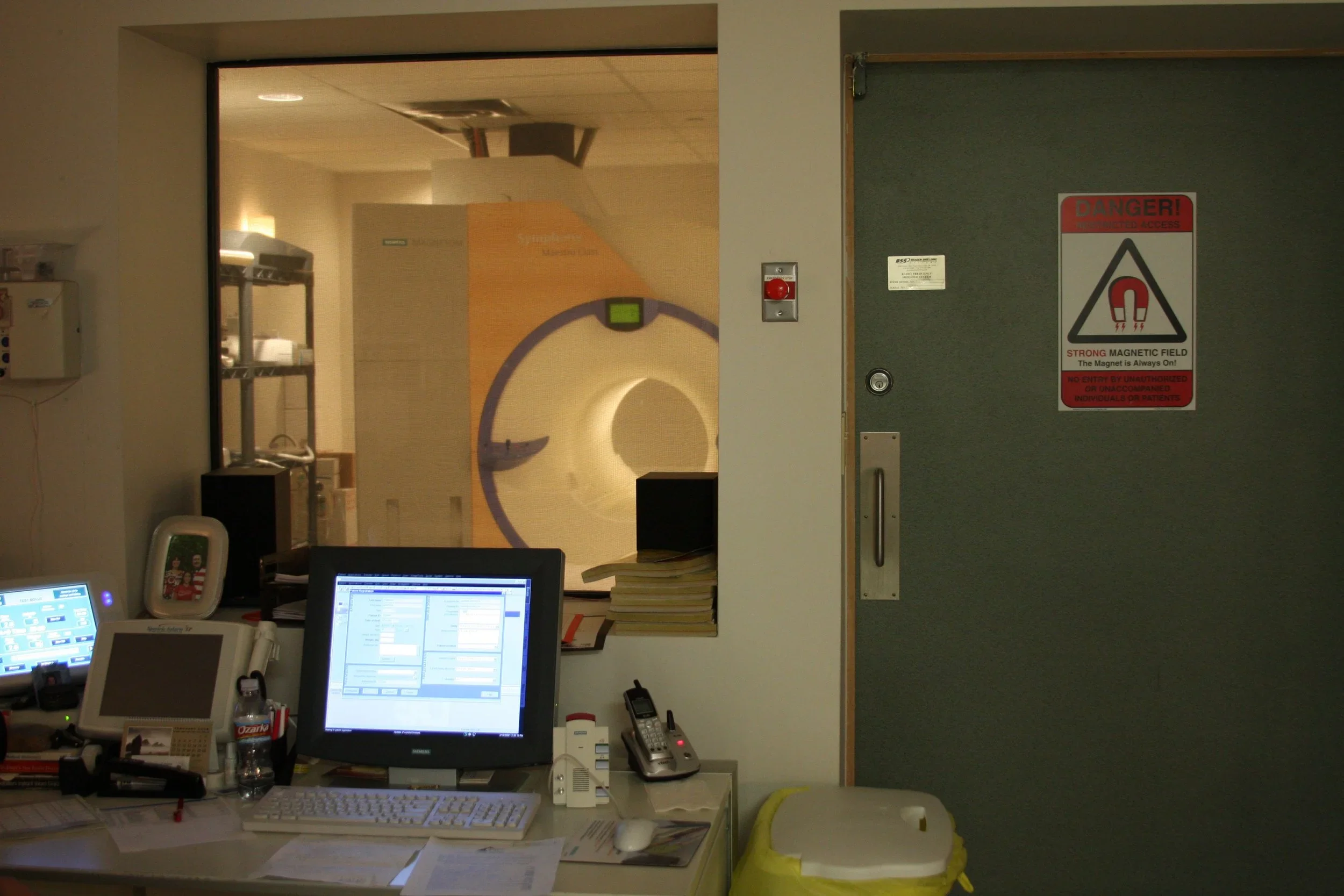 Medical imaging room with an MRI scanner visible through a window, and a workstation with computers, papers, and a water bottle.