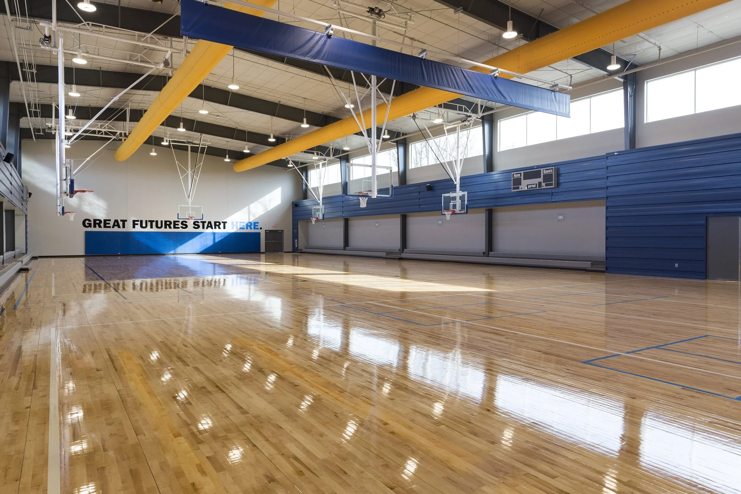 Empty indoor basketball court with polished wood flooring, blue and yellow gymnasium walls, multiple basketball hoops, and a motivational sign on the wall that reads 'GREAT FUTURES START HERE.'