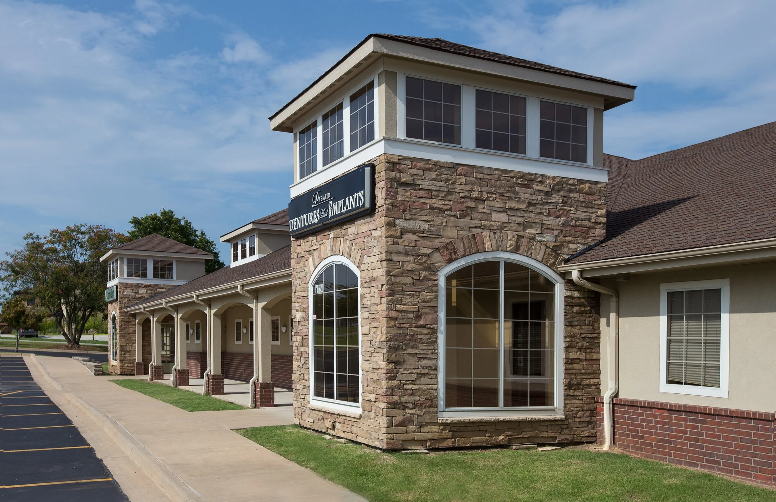 Exterior view of a building with stone and stucco walls, large arched windows, a sign for dentures and implants, and a parking lot in front.