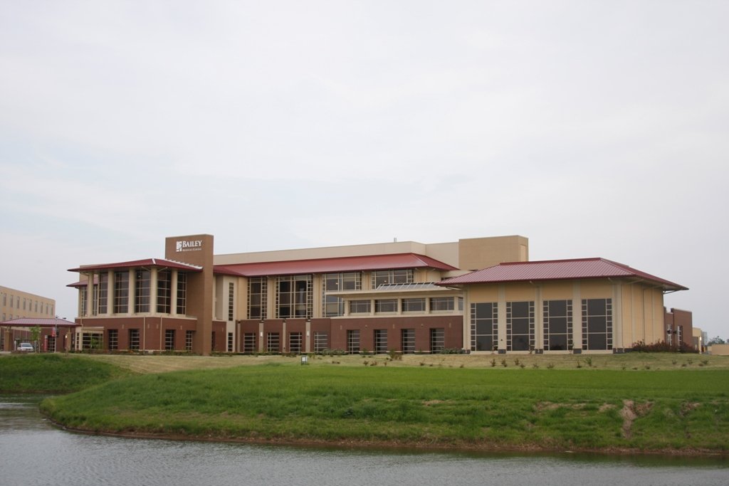 Modern multi-story office building with large windows and a red roof, located near a water body, with a lawn and landscaping in the foreground.