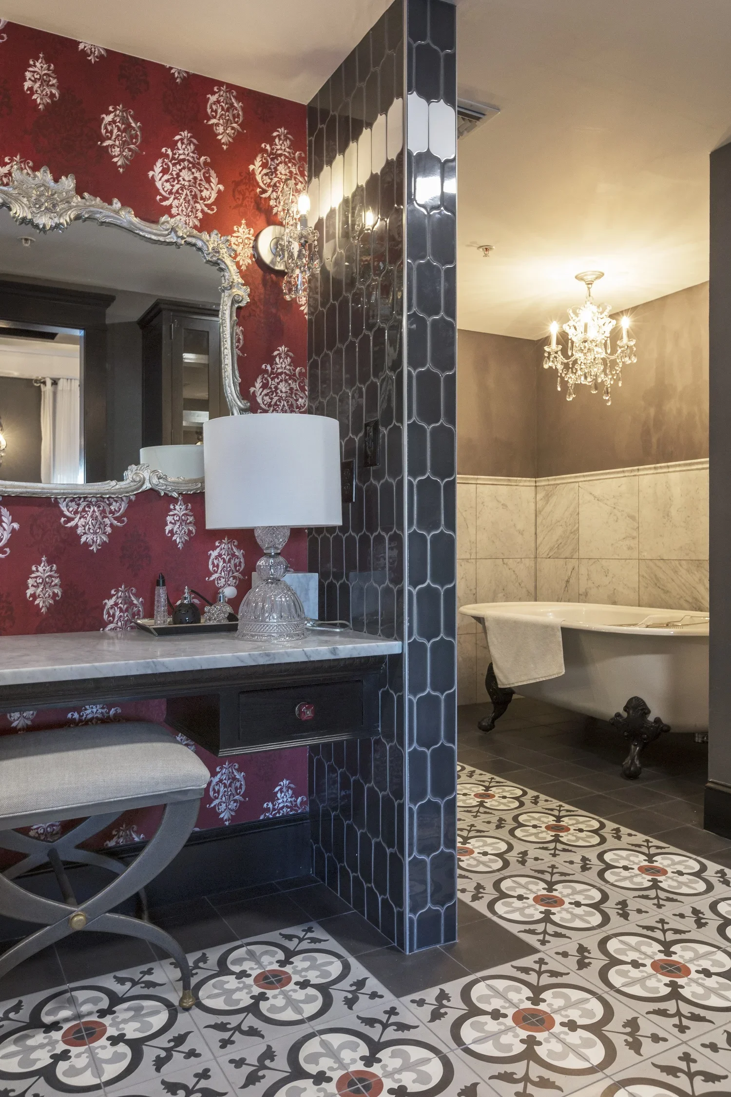 Decorative bathroom with red wallpaper, ornate mirror, white lamp, black vanity, patterned tile floor, and clawfoot bathtub with chandelier lighting.