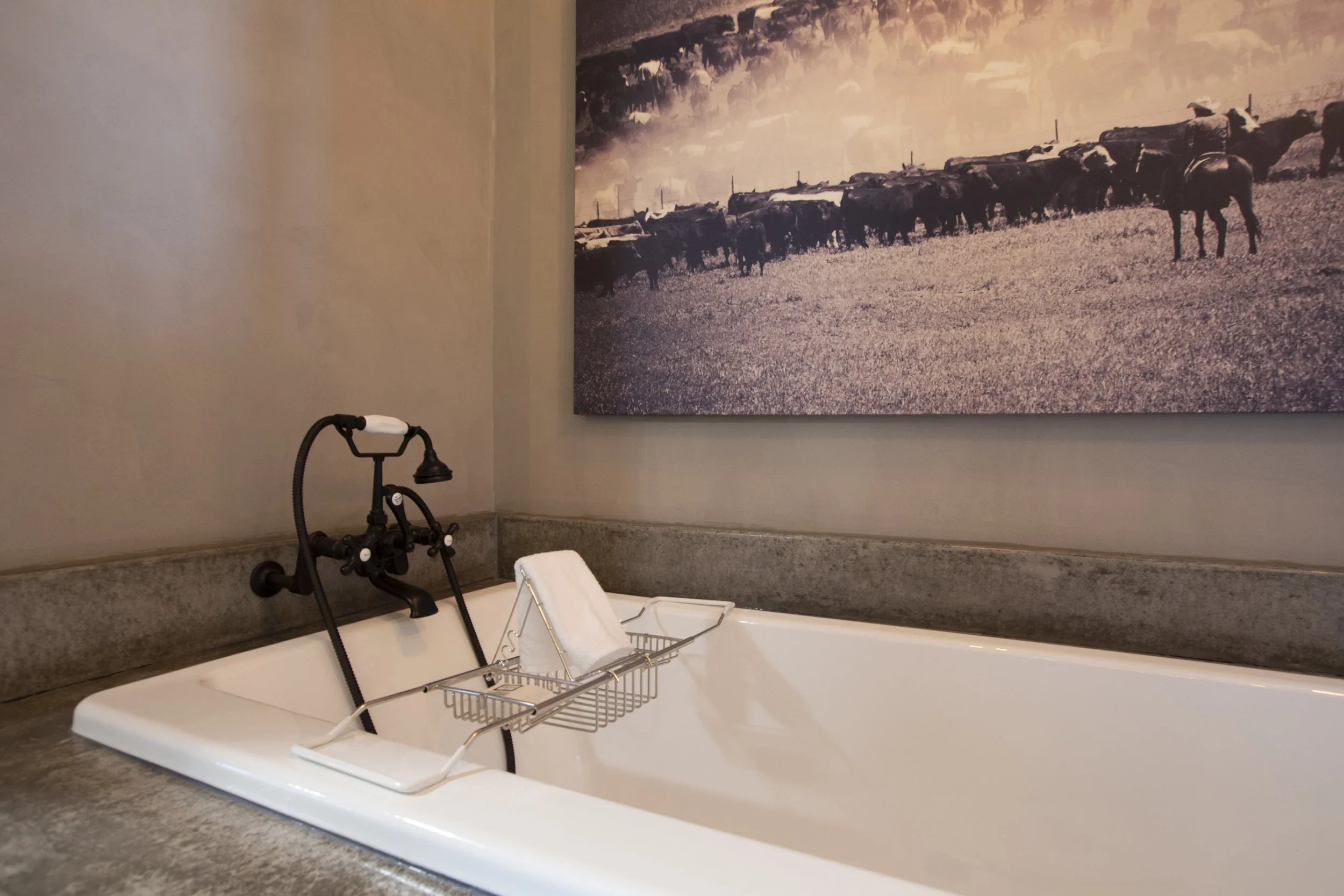A bathroom with a white bathtub, a black vintage-style faucet with a showerhead, a wire bath rack holding a folded white towel, and a large monochrome landscape painting of a herd of cows grazing on a field hung on the wall behind the bathtub.