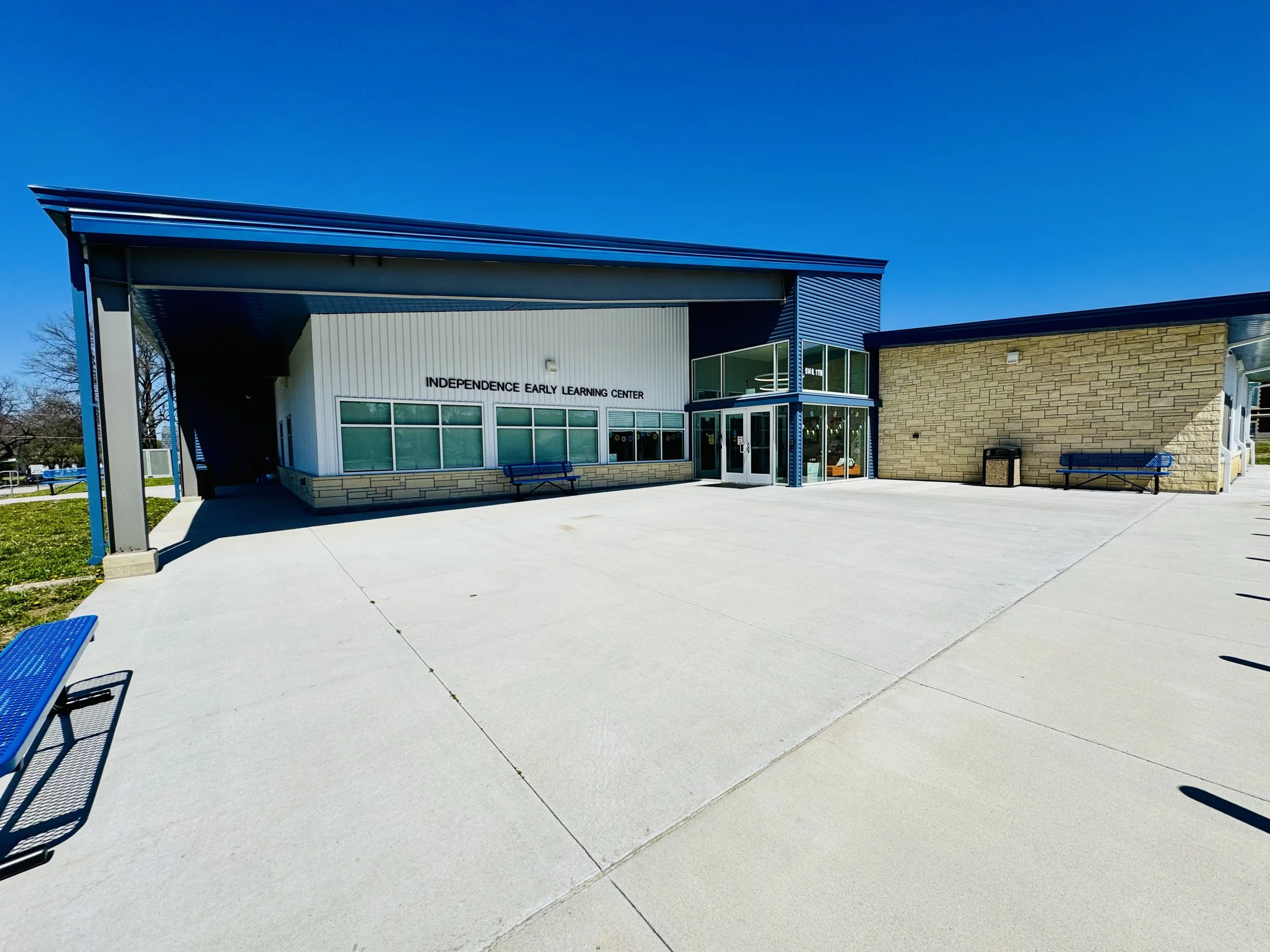 Exterior view of Independence Early Learning Center building with a wide concrete sidewalk, benches, and a clear blue sky.